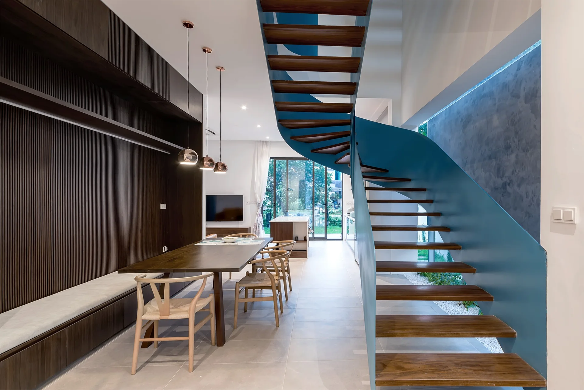 A modern open dining area features a large wood table, light wood chairs, dark fluted wood paneling, and a prominent curved teal staircase.