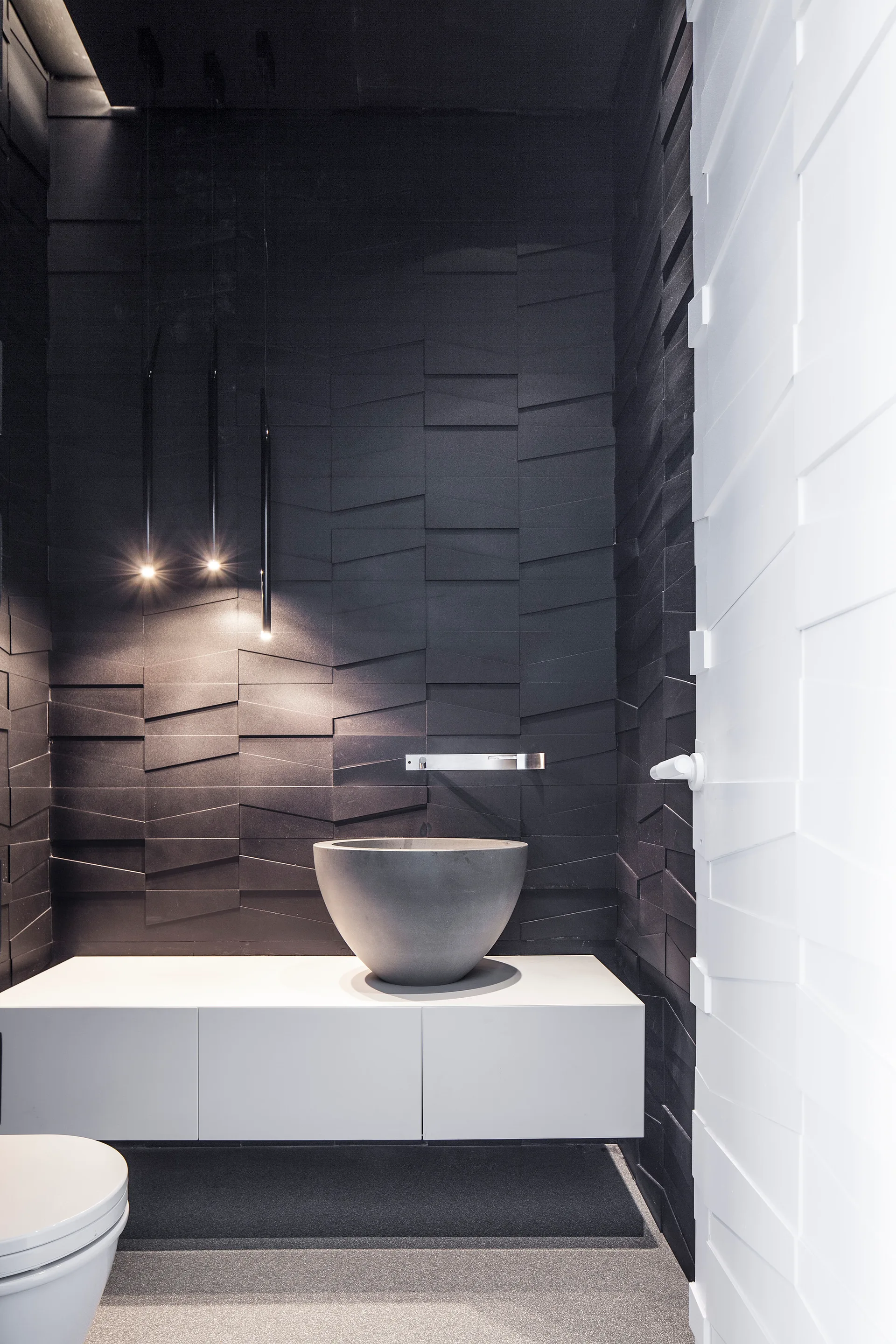 A modern powder room features a grey vessel sink on a white floating vanity against a black textured geometric tile wall illuminated by pendant lights.