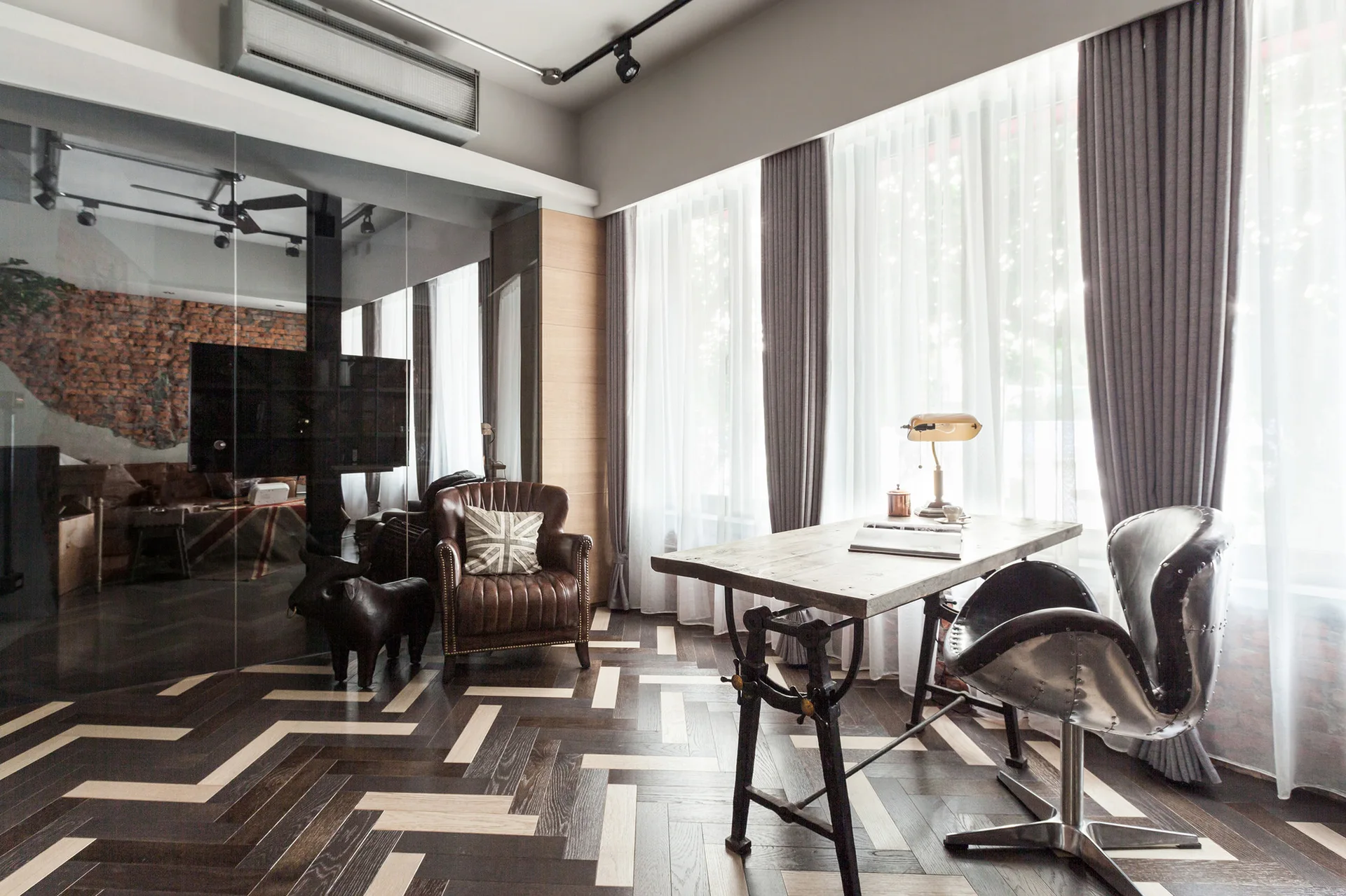 An industrial home office features a wood desk, a metal swivel chair, a brown leather armchair, and dark herringbone wood floors next to a glass wall.