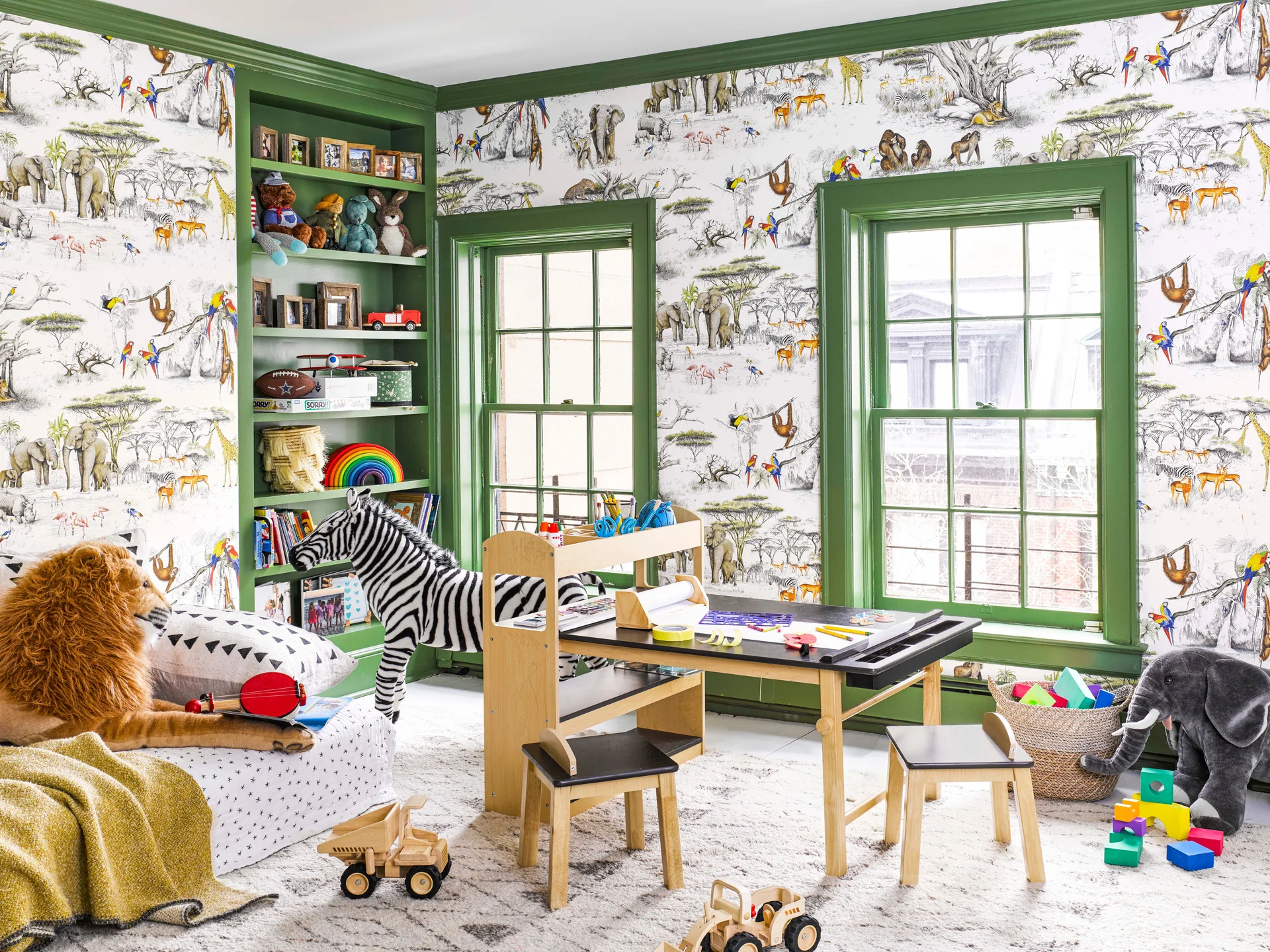 A kids playroom features safari themed wallpaper, green painted window trim and built in bookshelves, and a wooden activity table.