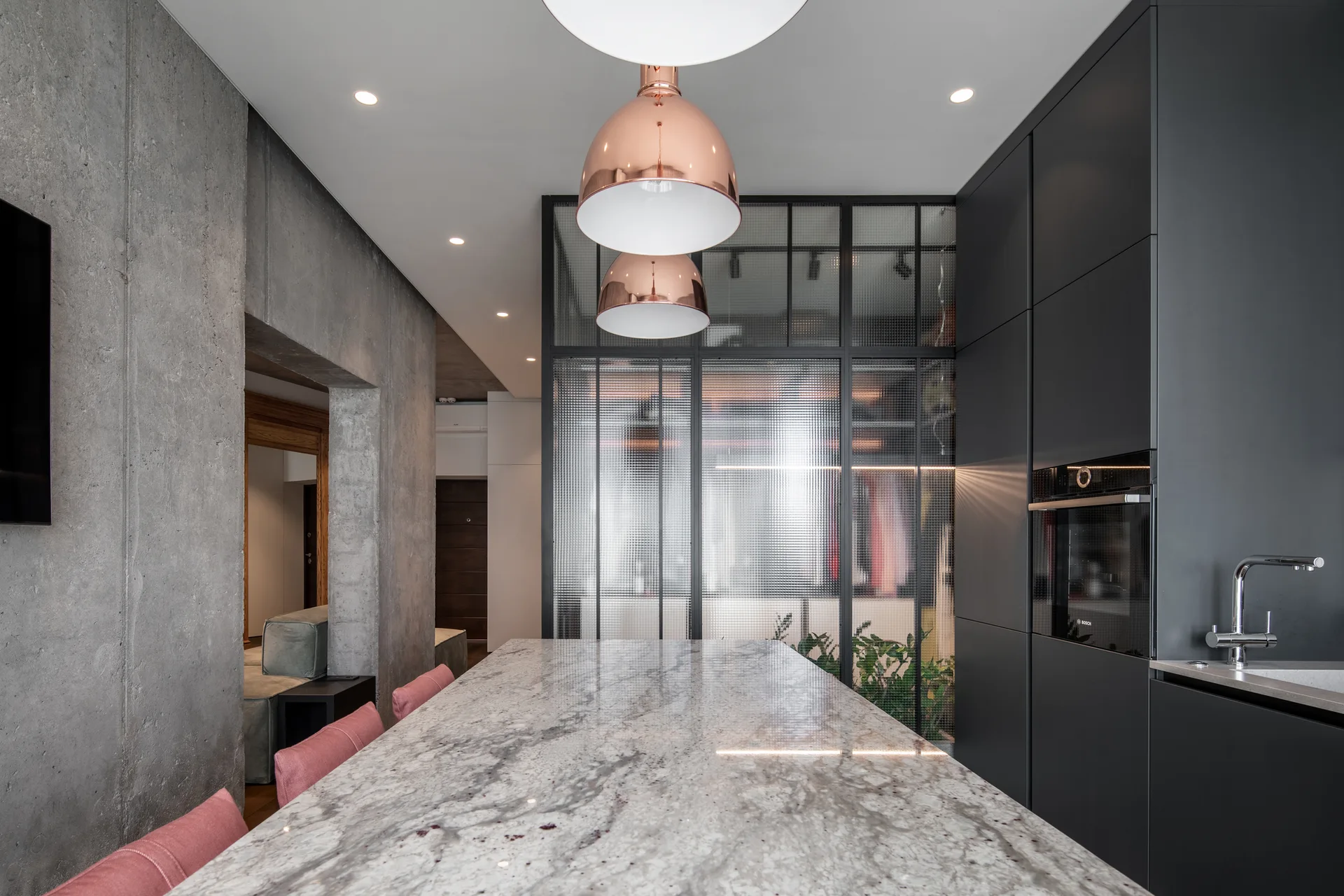 A modern kitchen features a grey stone island with pink chairs, copper dome pendant lights, matte black cabinets, and a textured glass partition.