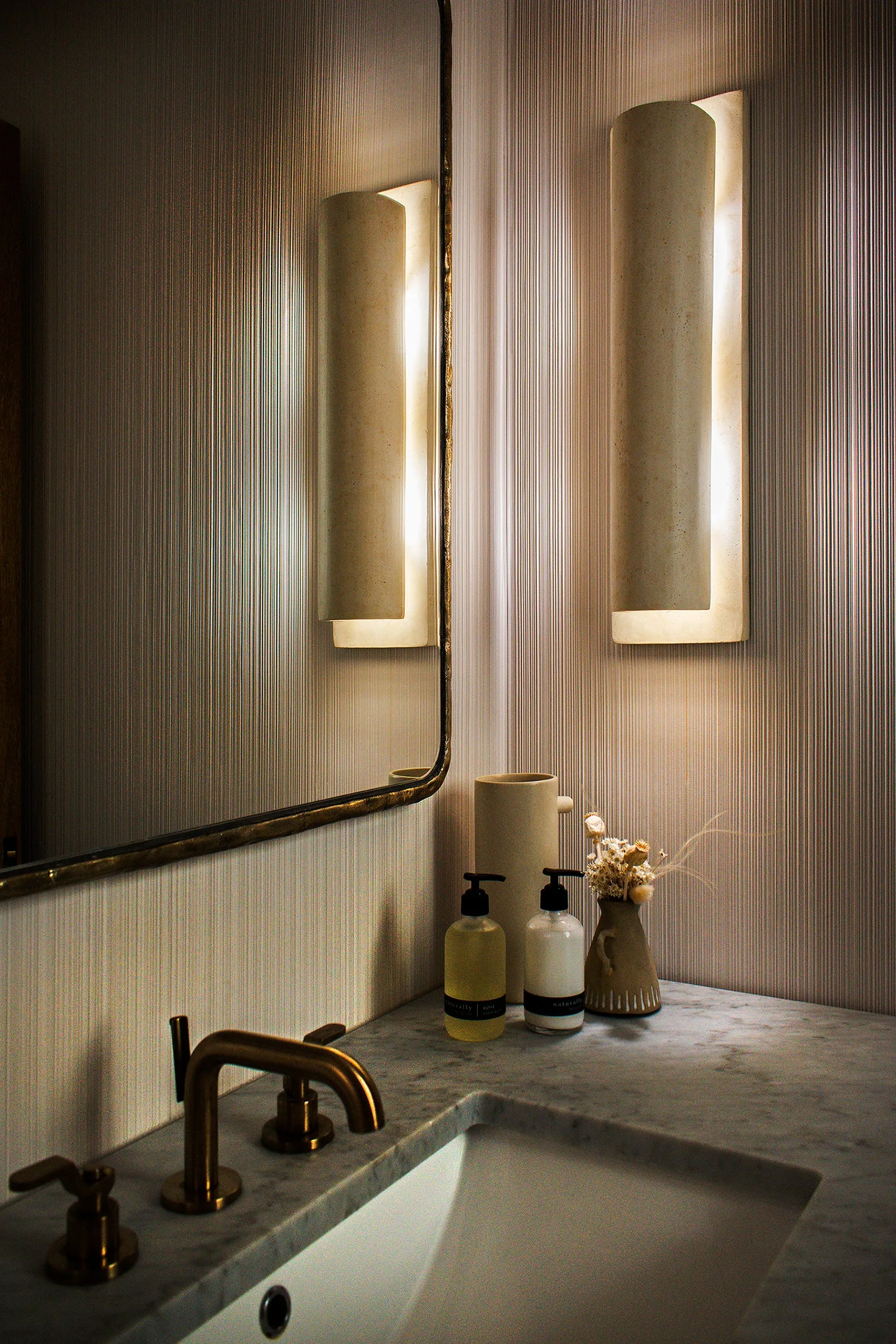 A powder room vanity features a grey stone countertop, a brass faucet, a framed mirror, and a glowing stone wall sconce against textured wallpaper.