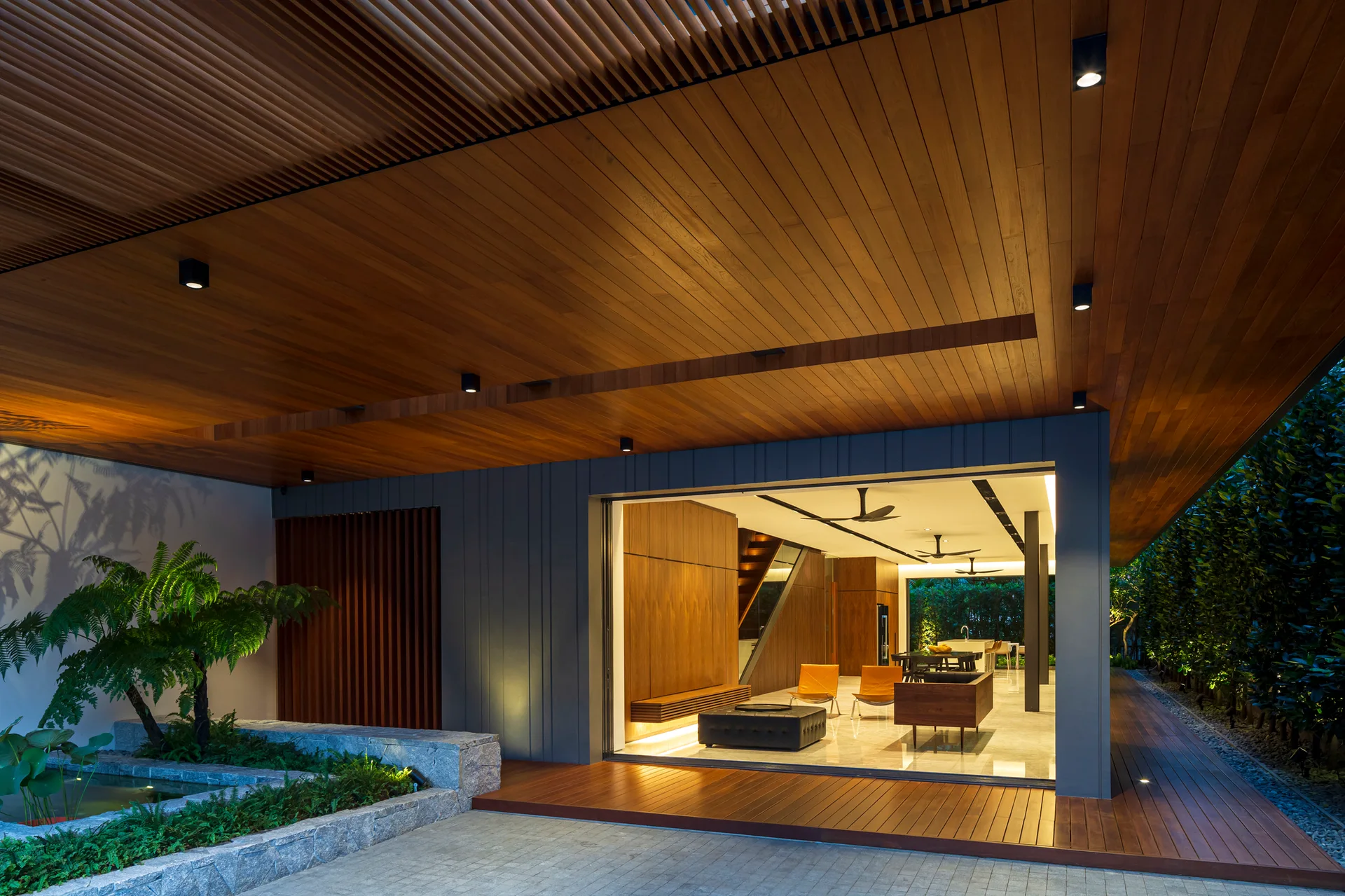 A modern covered patio features a rich wood slat ceiling, wooden decking, a landscaped stone pond, and a wide open doorway leading into a warm interior lounge.