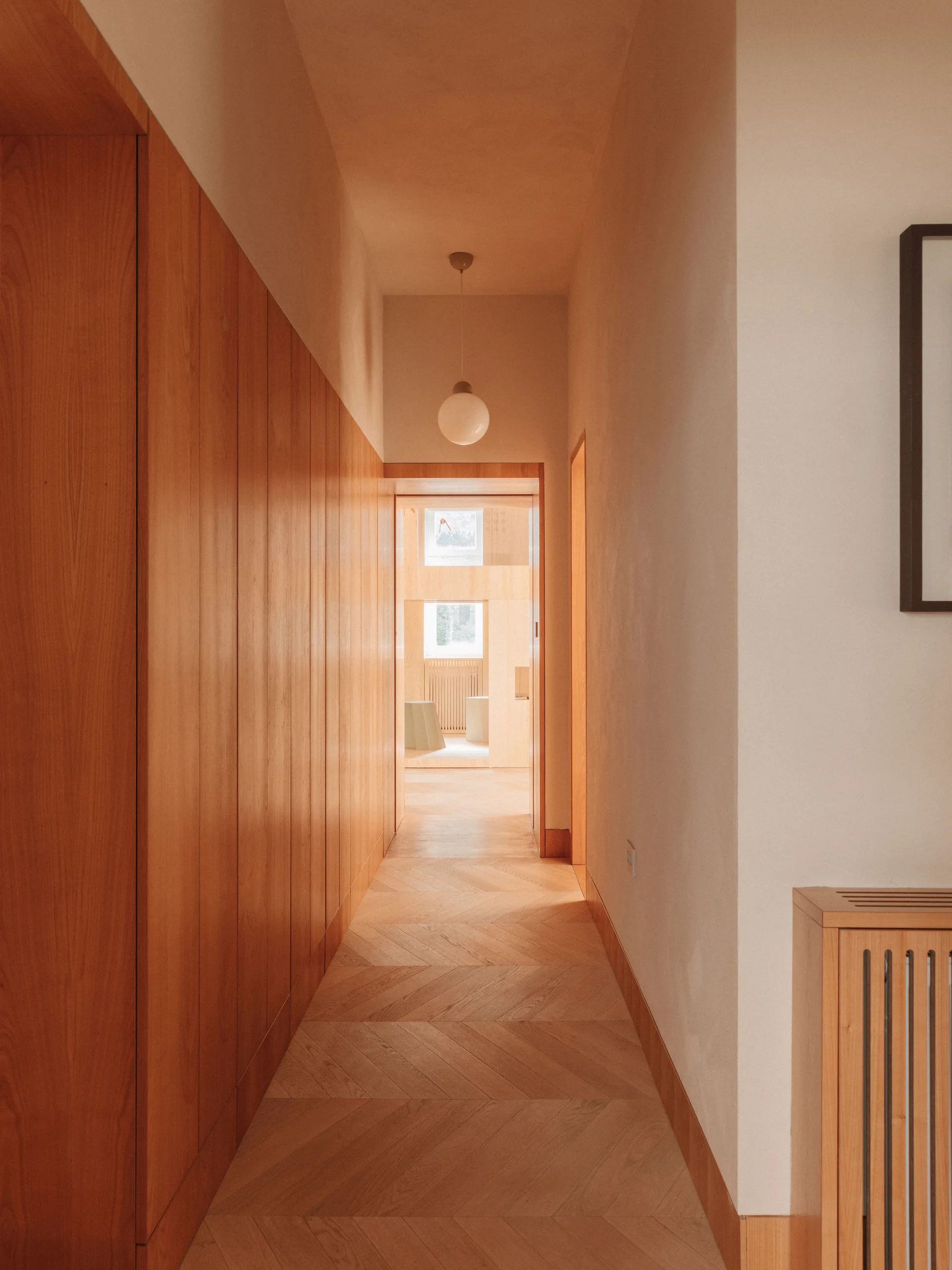 A narrow hallway featuring vertical wood wall paneling, a light wood herringbone floor, and a glowing globe pendant light.