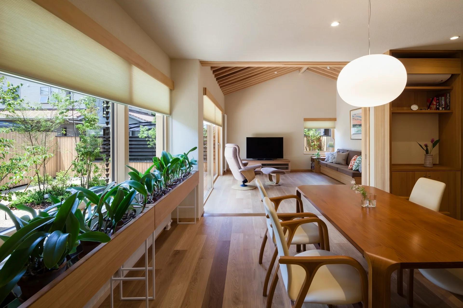 An open-concept dining and living space features light wood flooring, a long wooden planter along large windows, and a sloped ceiling with exposed beams.