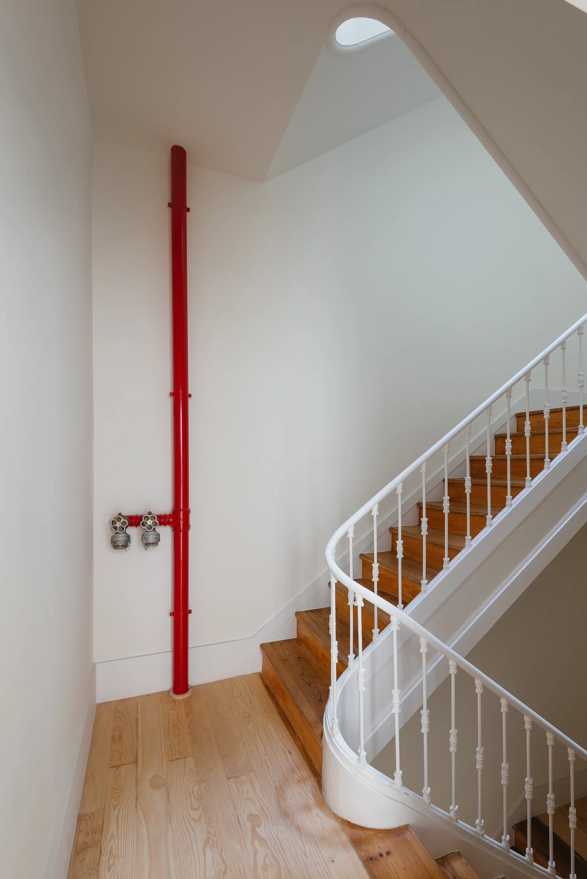 A curved wooden staircase with white railings winds through a bright white stairwell alongside a prominent red vertical fire pipe.
