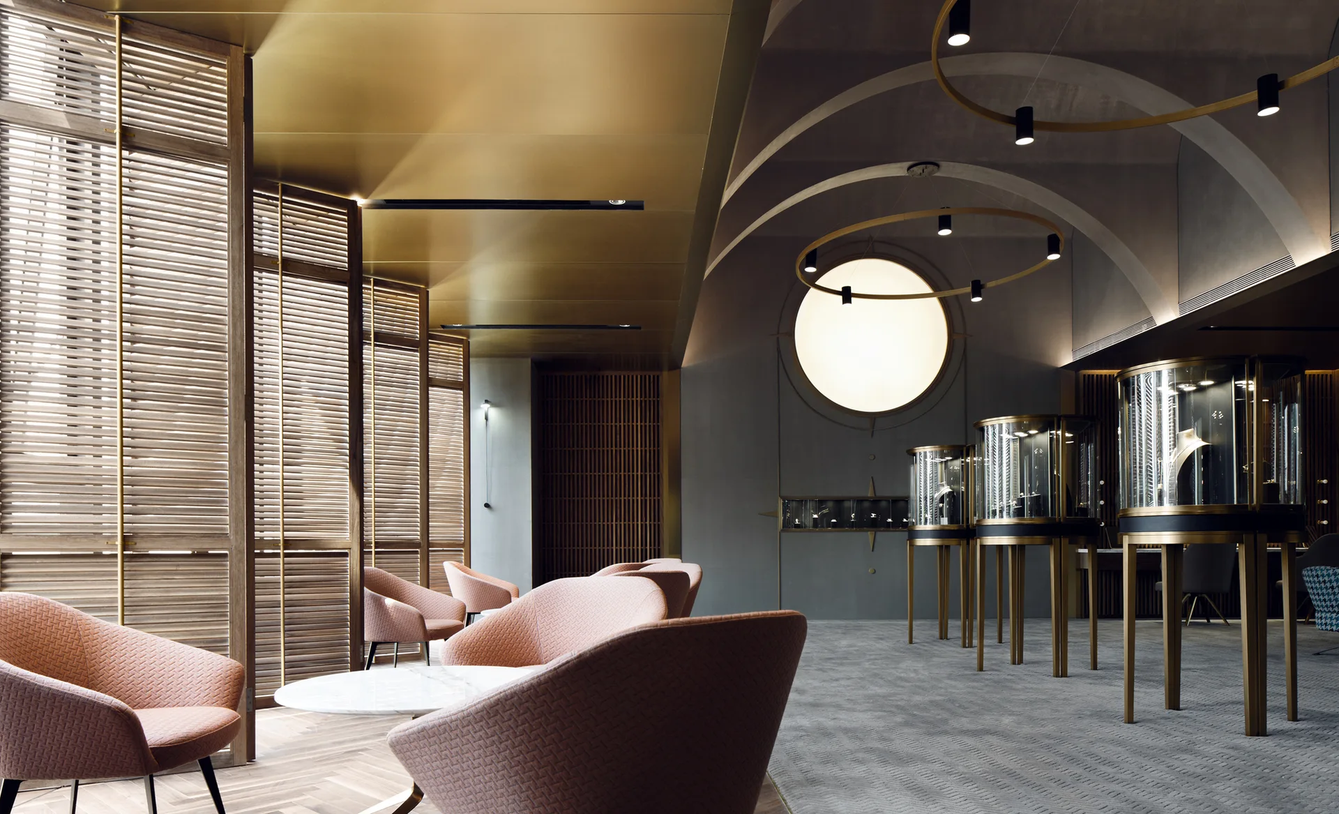 A modern lounge features dusty pink armchairs, tall wooden louvered shutters, circular glass jewelry displays, and a vaulted ceiling with gold accents.