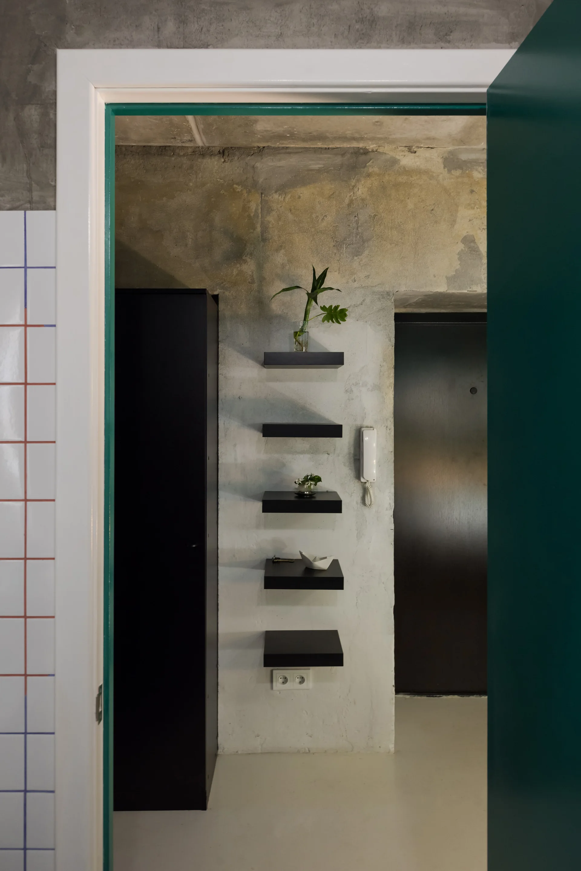 An entryway seen through a doorway featuring a raw concrete wall with five black floating shelves and a tall black cabinet.