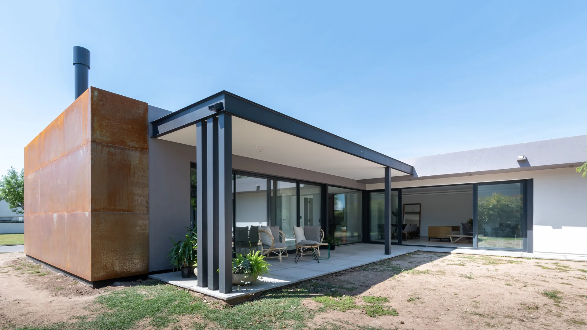 A modern home features a covered concrete patio with sliding glass doors, dark metal pillars, and a large weathered steel chimney structure.