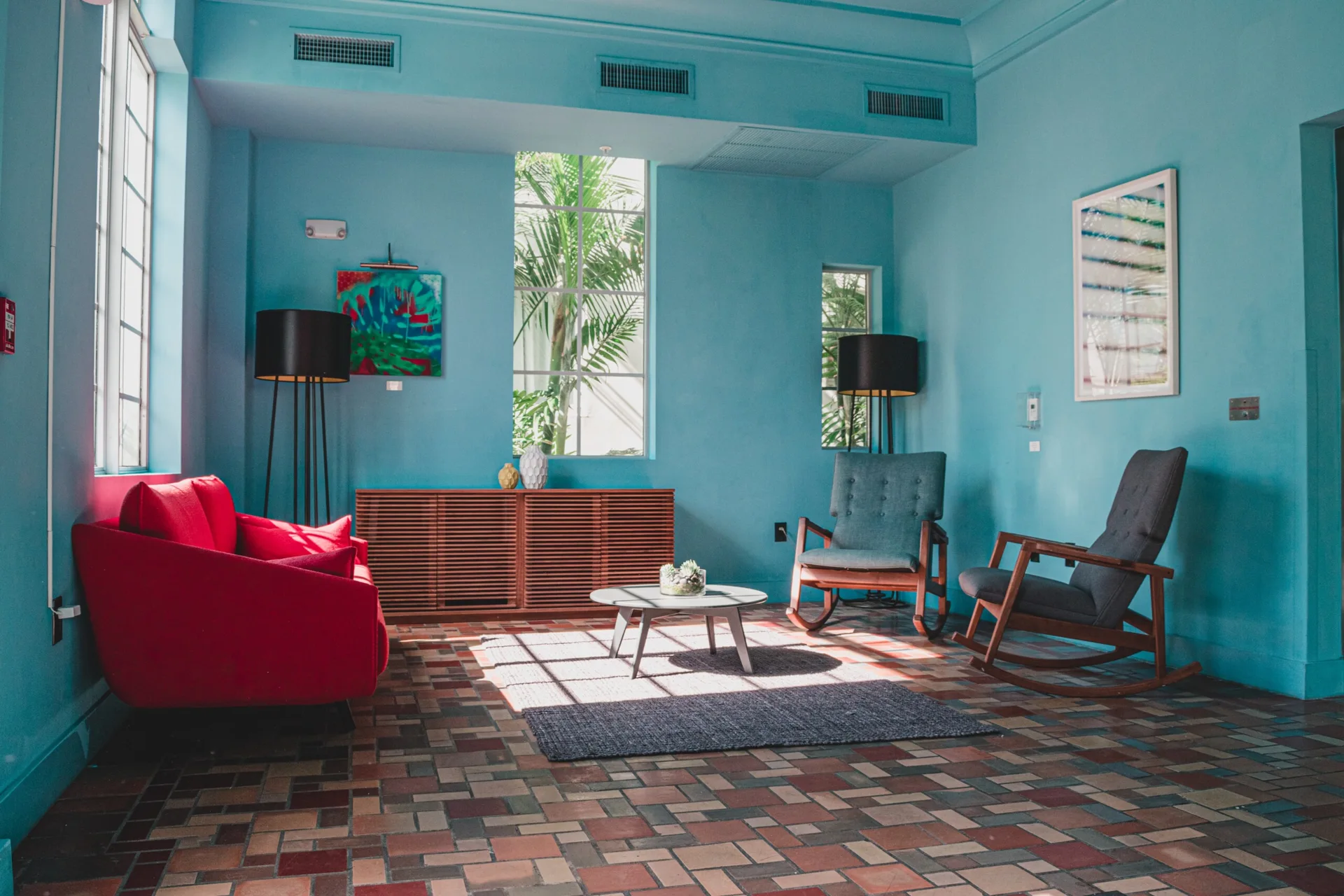 A bright blue living room features a bold red sofa, two grey rocking chairs, a slatted wooden cabinet, and a multi-colored tiled floor.