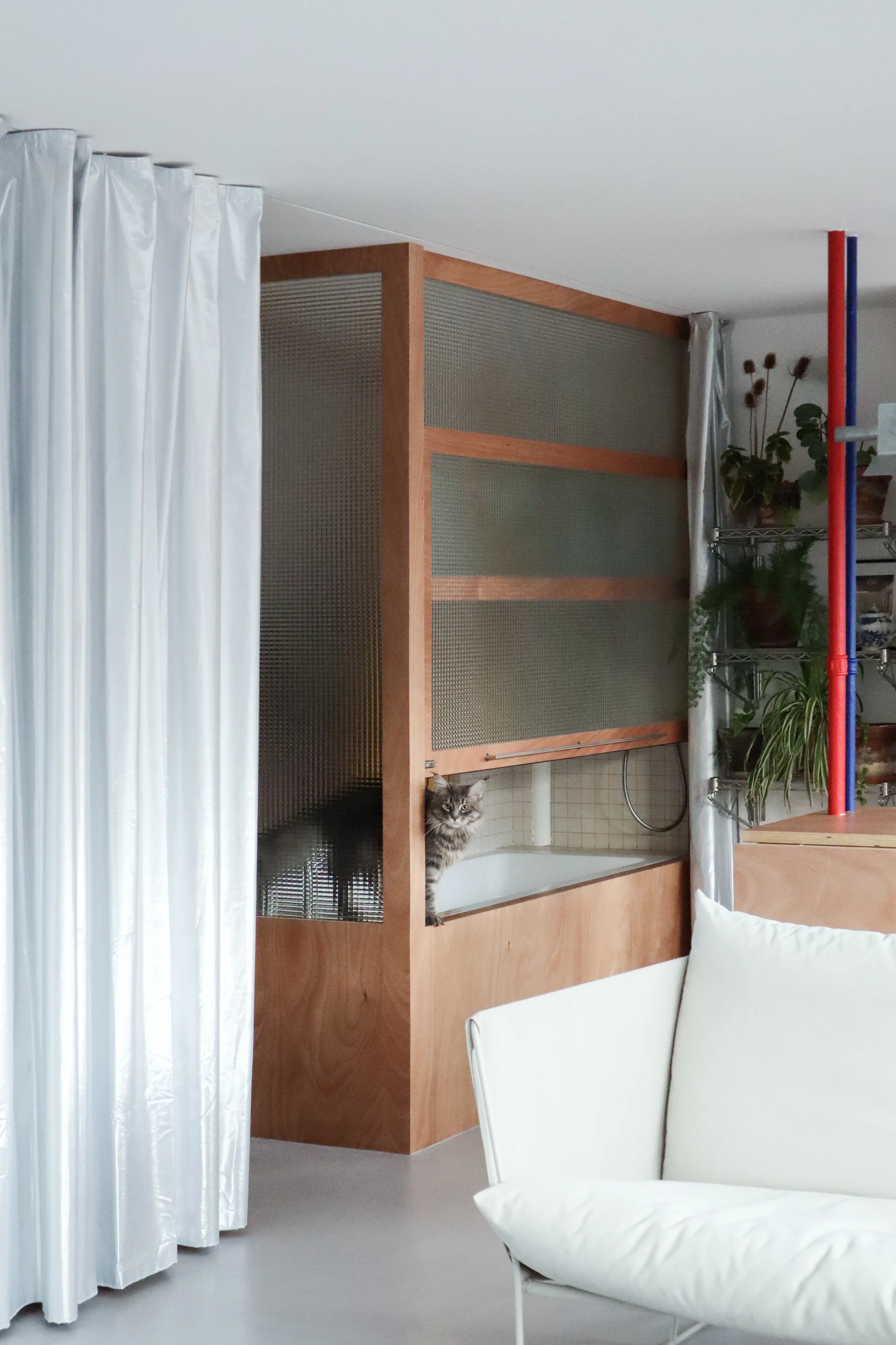 A wood-framed textured glass partition encloses a white bathtub next to a living area with a white sofa and decorative plants.