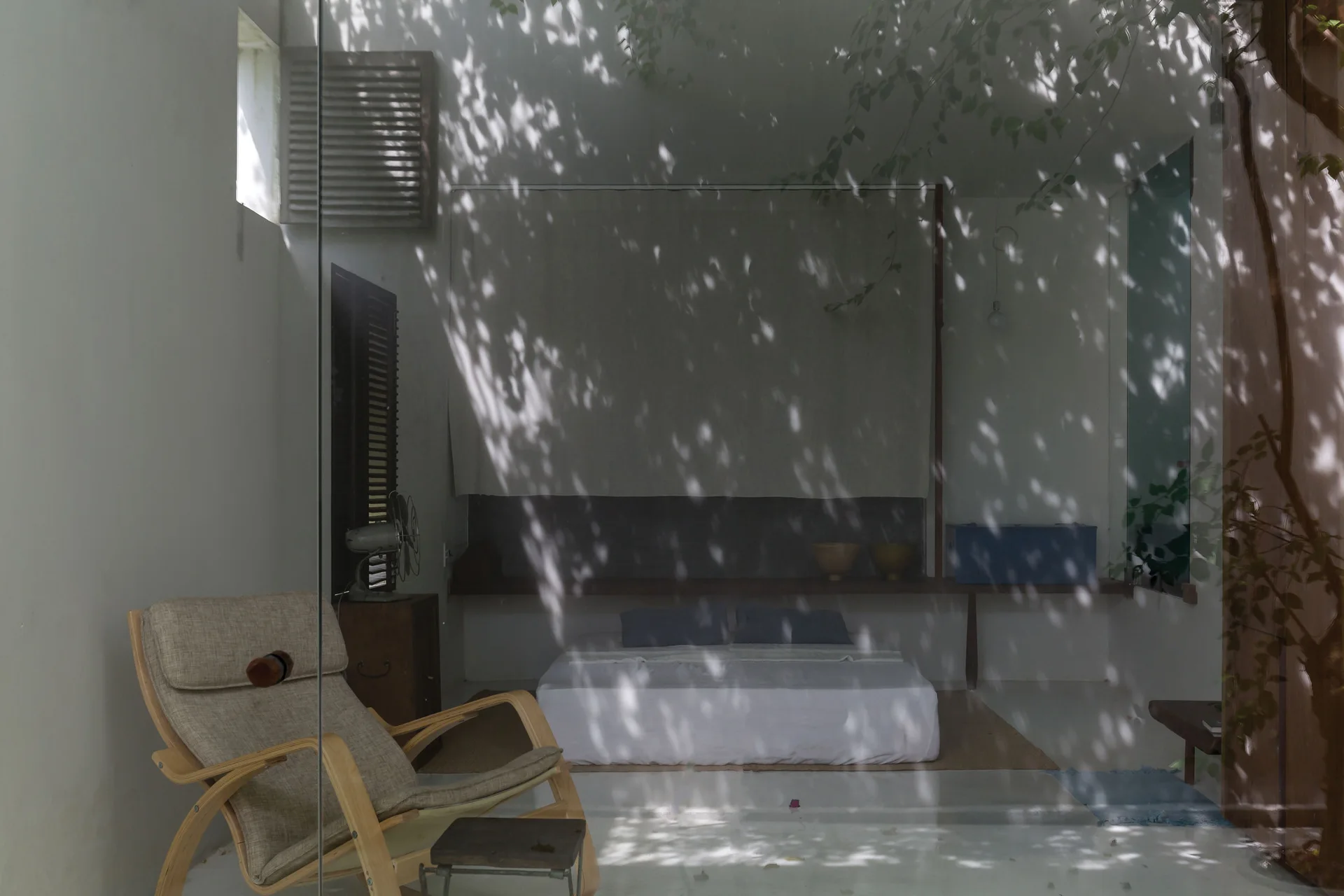 A minimalist bedroom with a low platform bed and wooden lounge chair, viewed through a glass panel reflecting tree shadows on white walls.