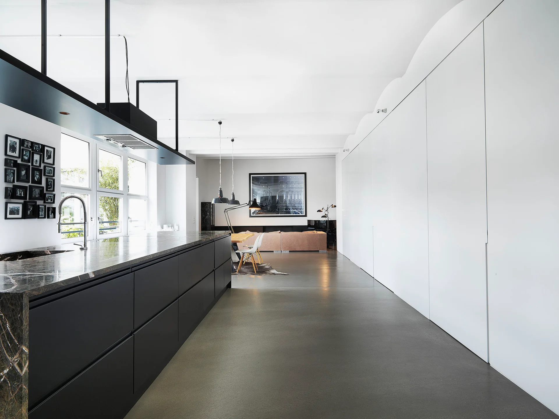 A long matte black kitchen island with a dark stone countertop sits opposite a full wall of seamless white cabinetry on a smooth grey floor.