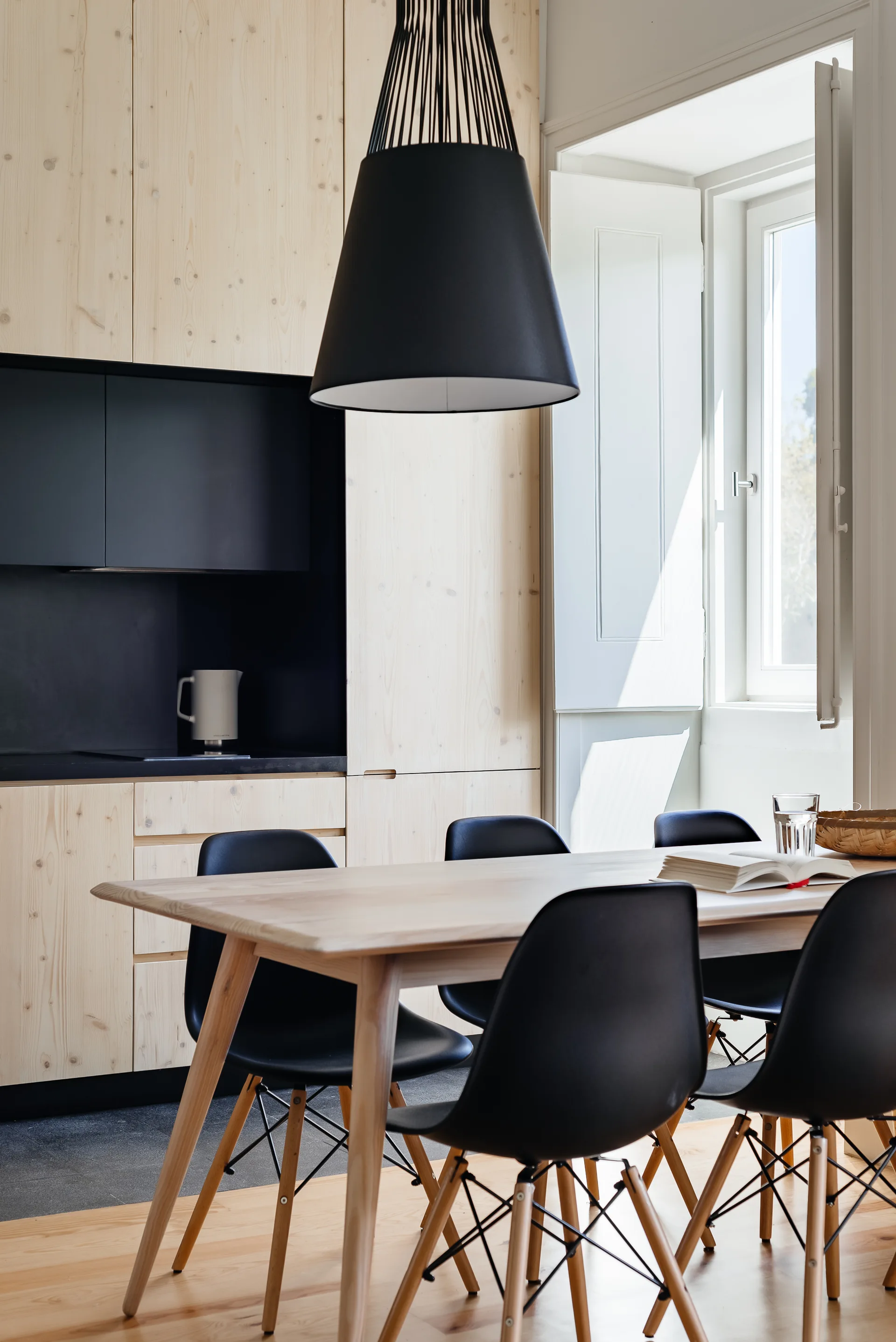 A light wood dining table with black chairs sits next to matching wood kitchen cabinets and a large black pendant light hanging above.