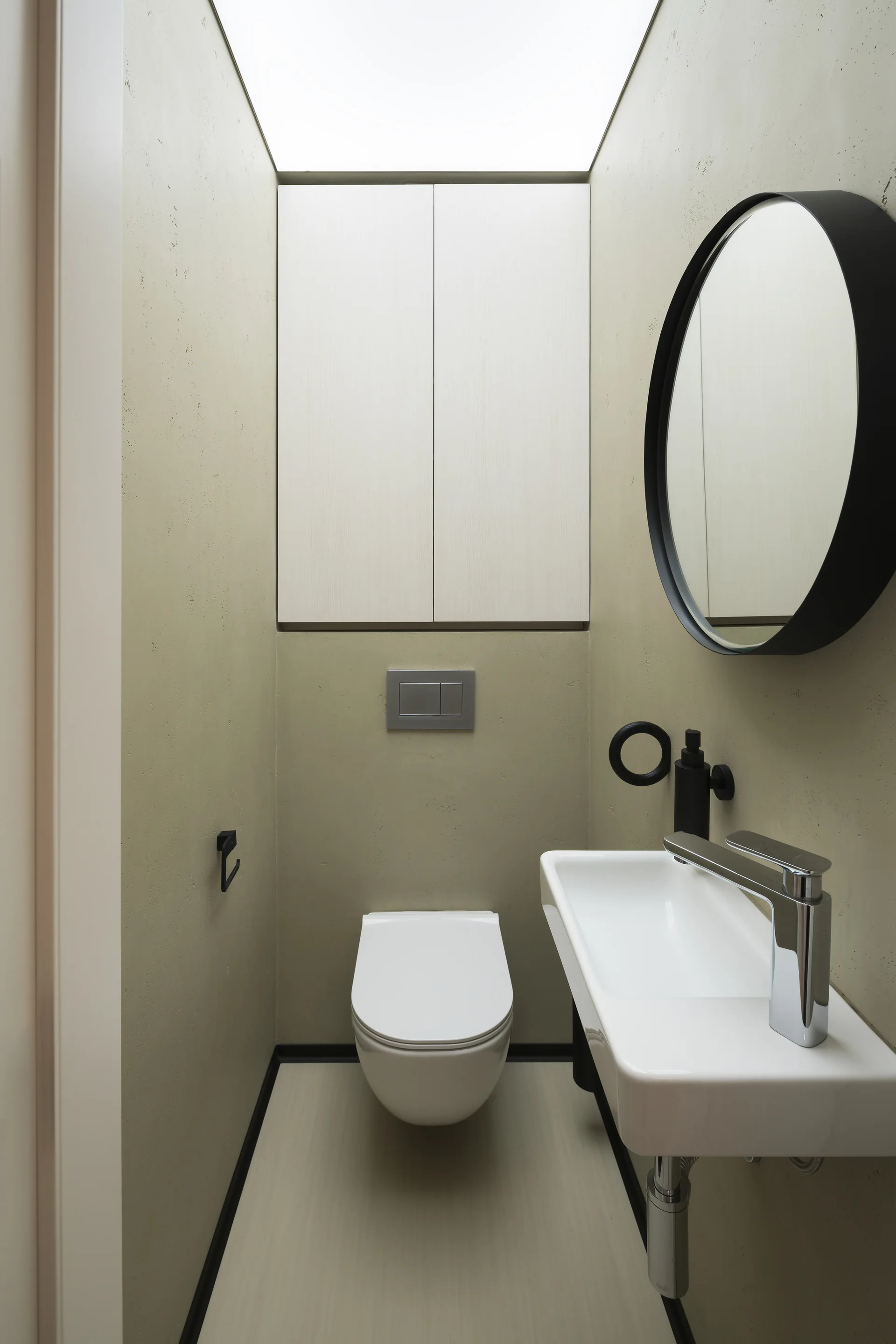 A minimalist powder room features sage green walls, a wall-hung toilet, a narrow white wall-mounted sink, and a large round black-framed mirror.