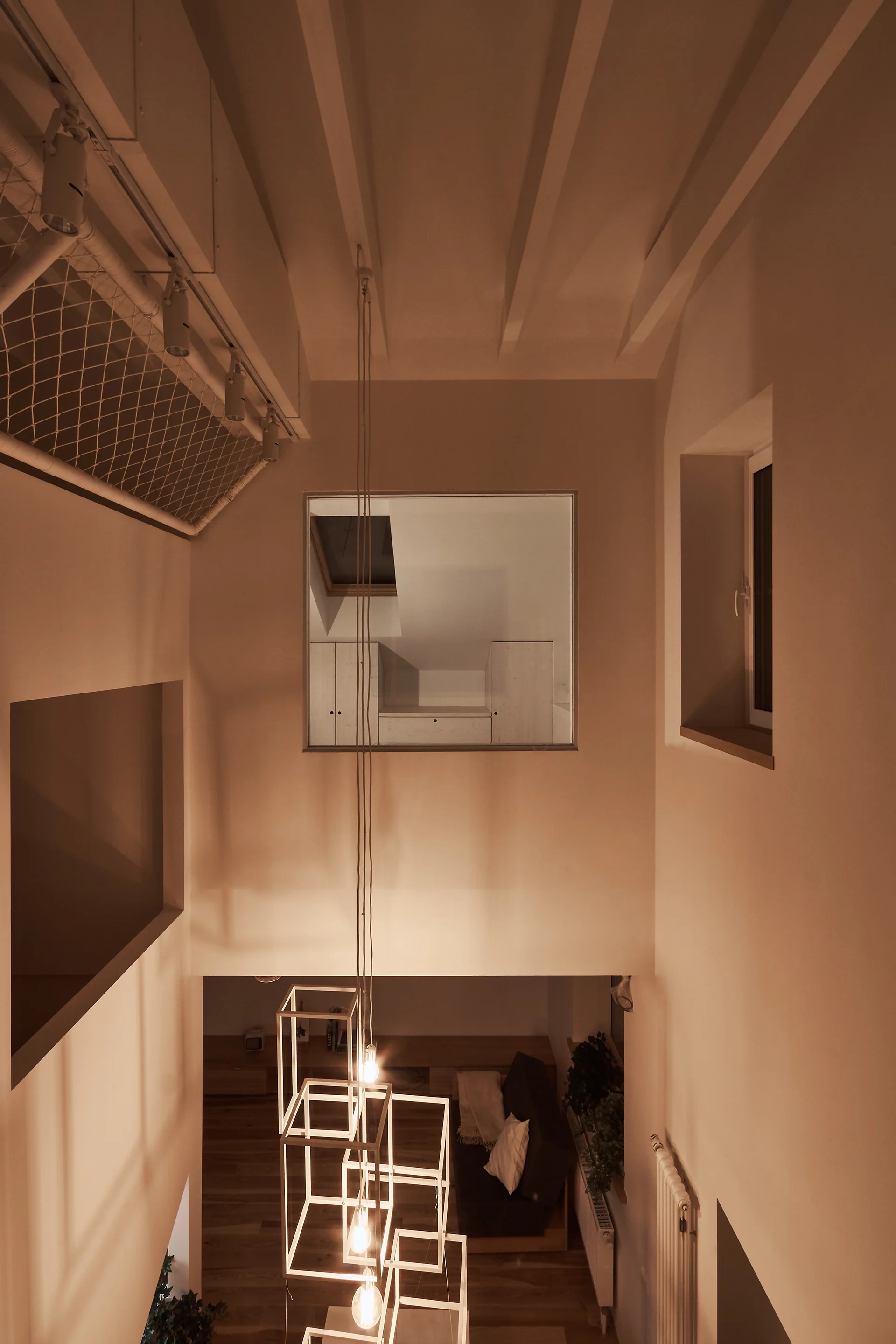 Looking down into a warm multi-level room with white walls, exposed ceiling beams, and a long geometric pendant light hanging above a sofa below.