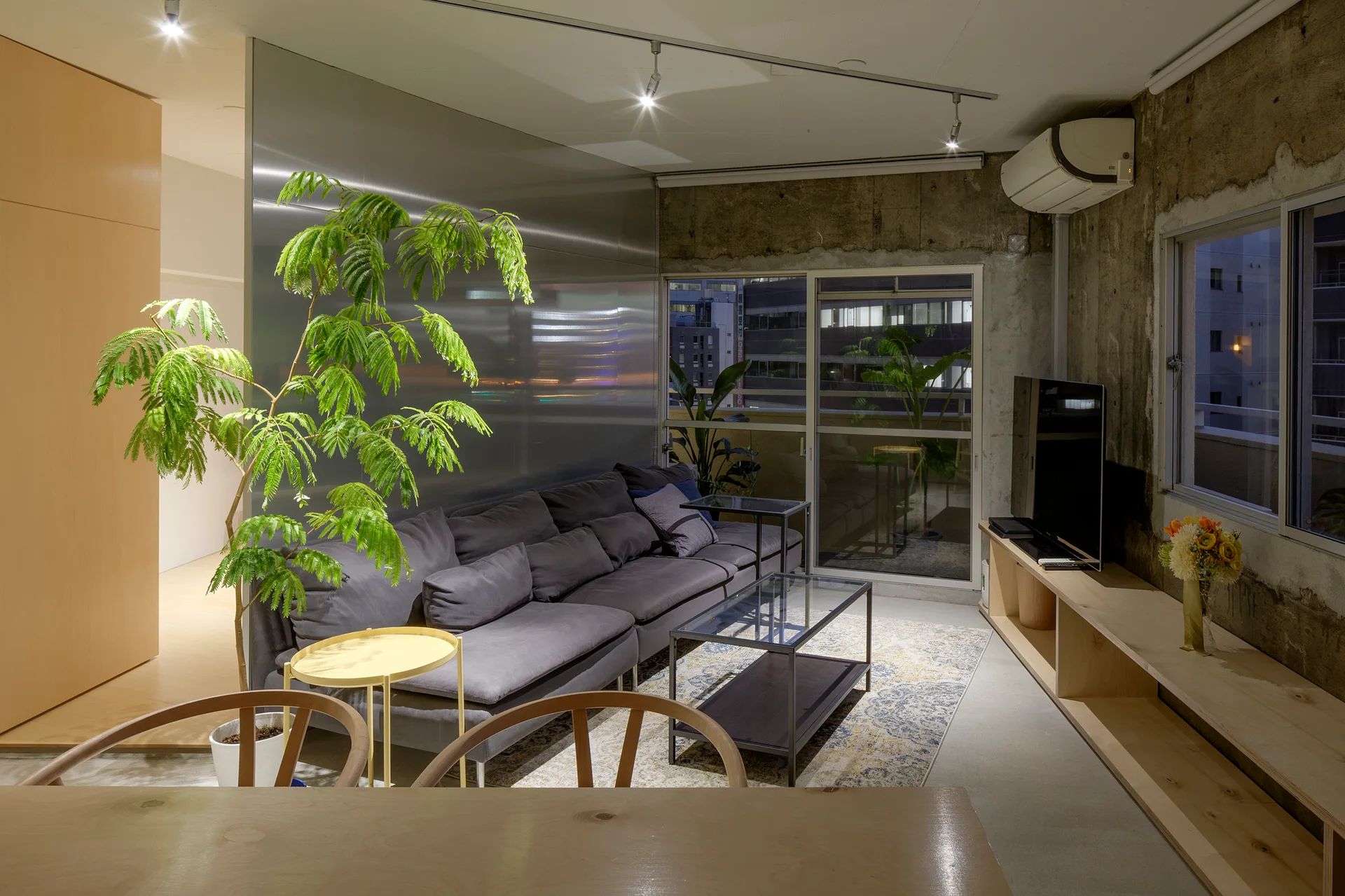 A grey modular sofa sits next to a metal partition wall in an industrial living room with exposed concrete and a long wooden media unit.