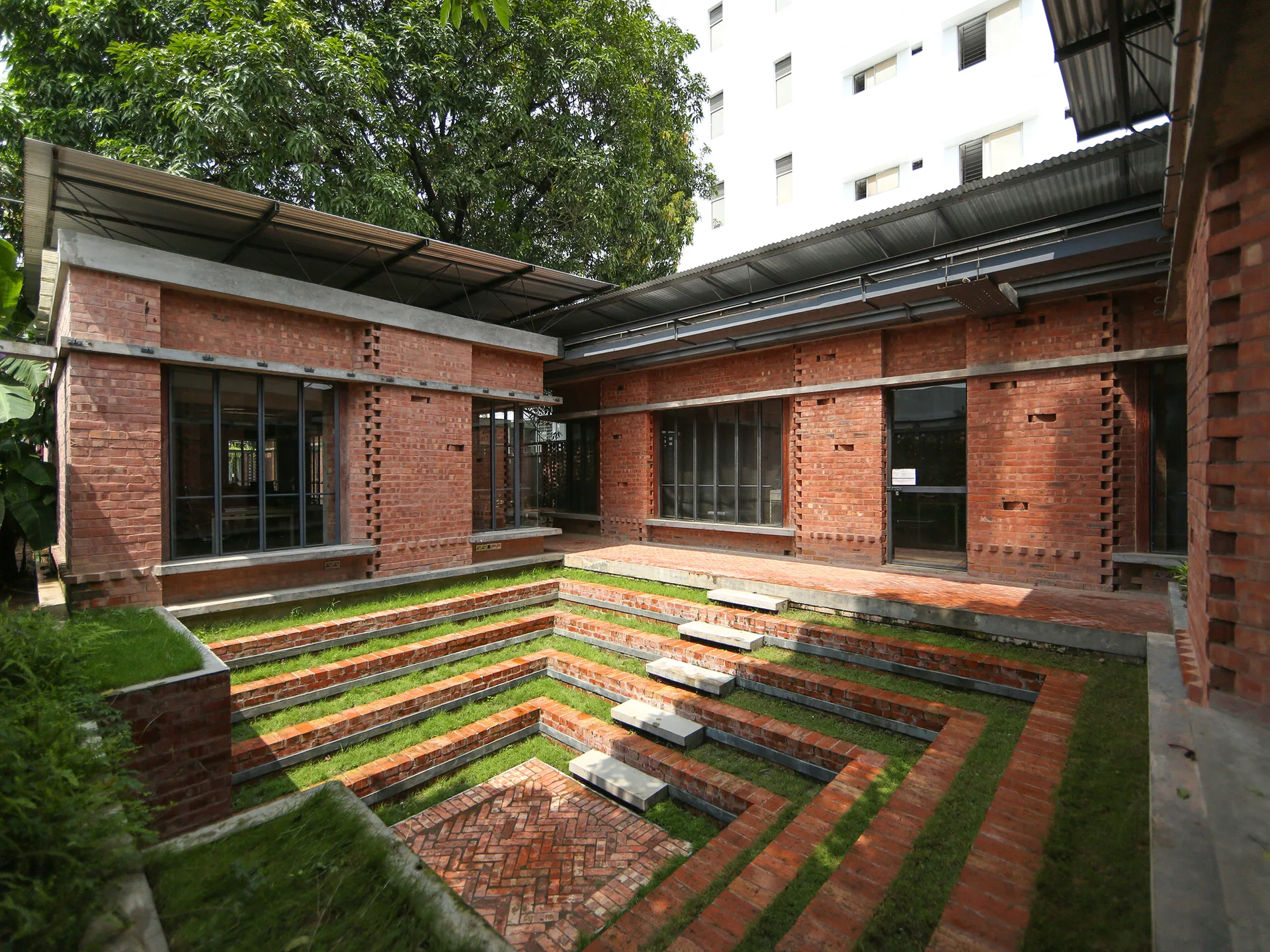 A sunken brick courtyard features stepped terracotta seating with embedded grass paths, surrounded by exposed brick walls and black-framed windows.