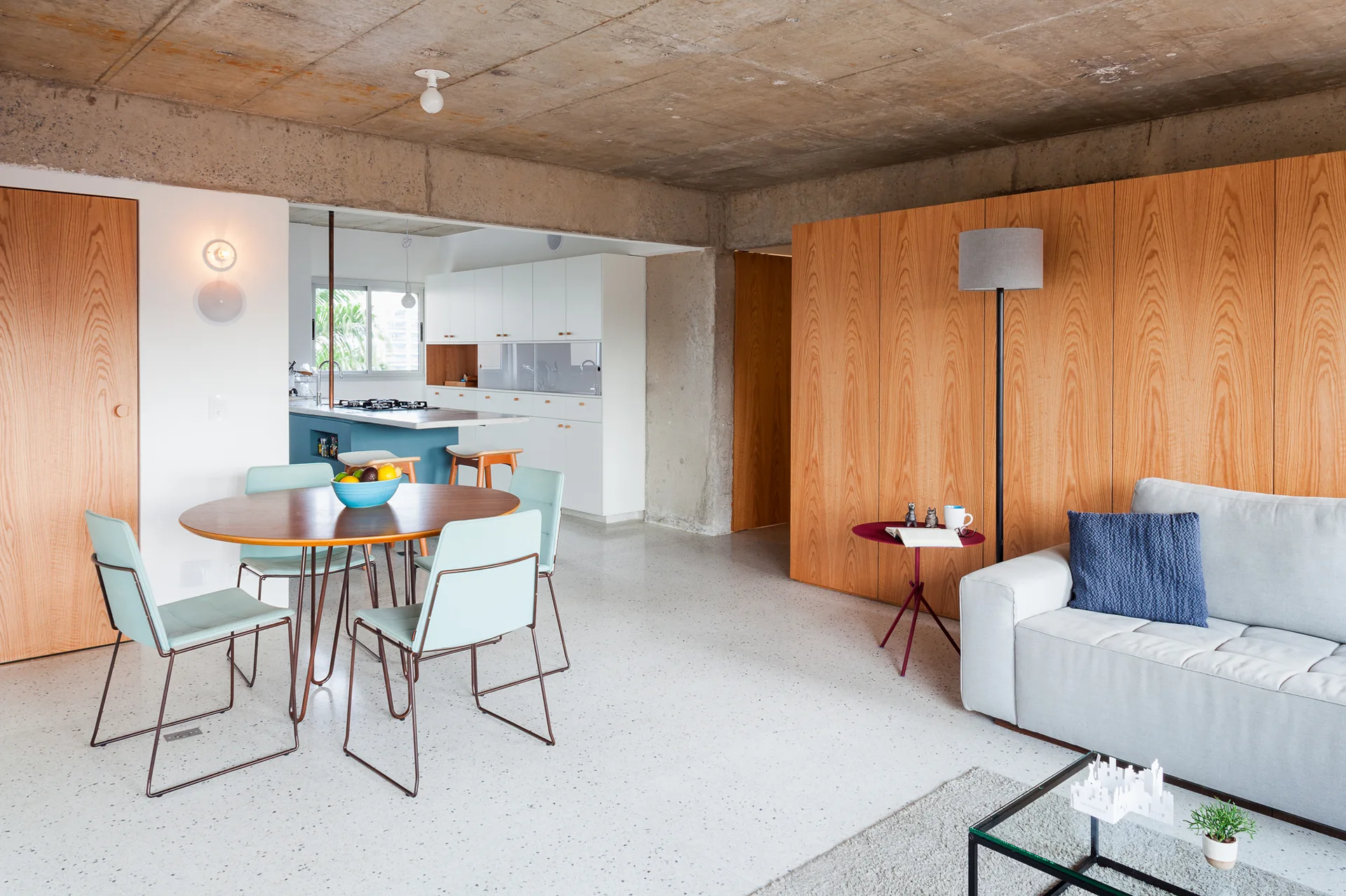 A modern open living and dining area featuring a raw concrete ceiling, warm wood paneling, light blue dining chairs, and a grey sofa.