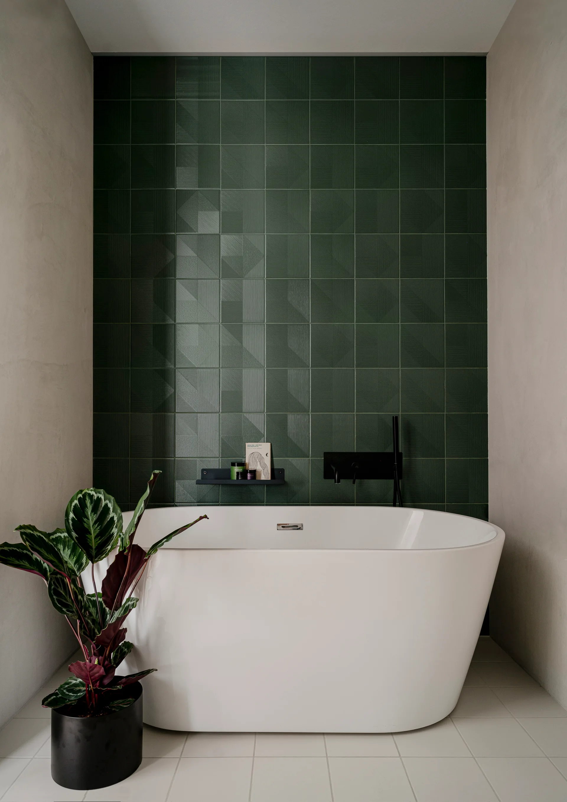 A white freestanding bathtub sits against a dark green textured tile wall next to a potted plant in a black planter.