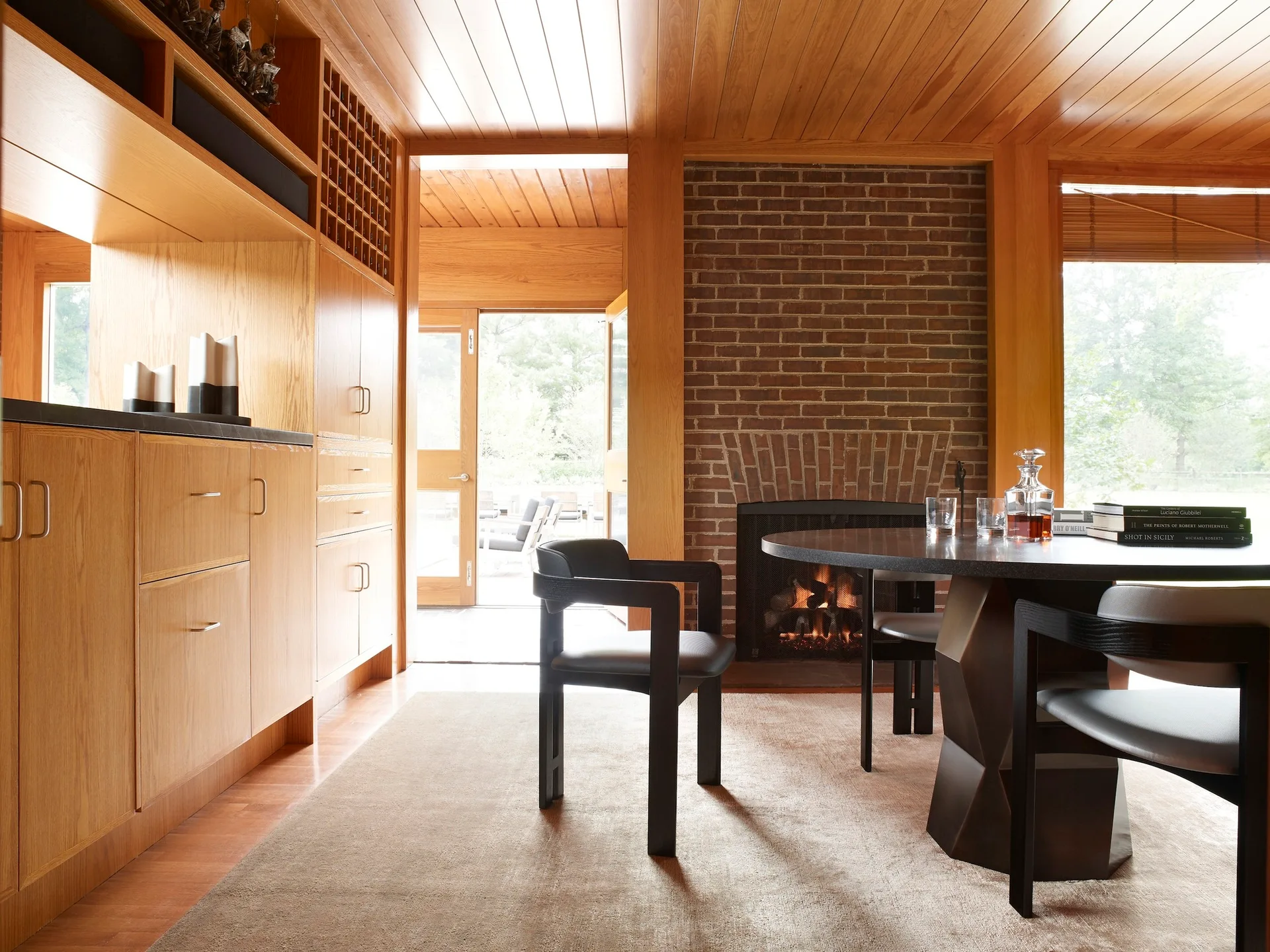 A mid-century modern dining room featuring a black circular table with chairs, warm wood-paneled walls and ceiling, and a brick fireplace.