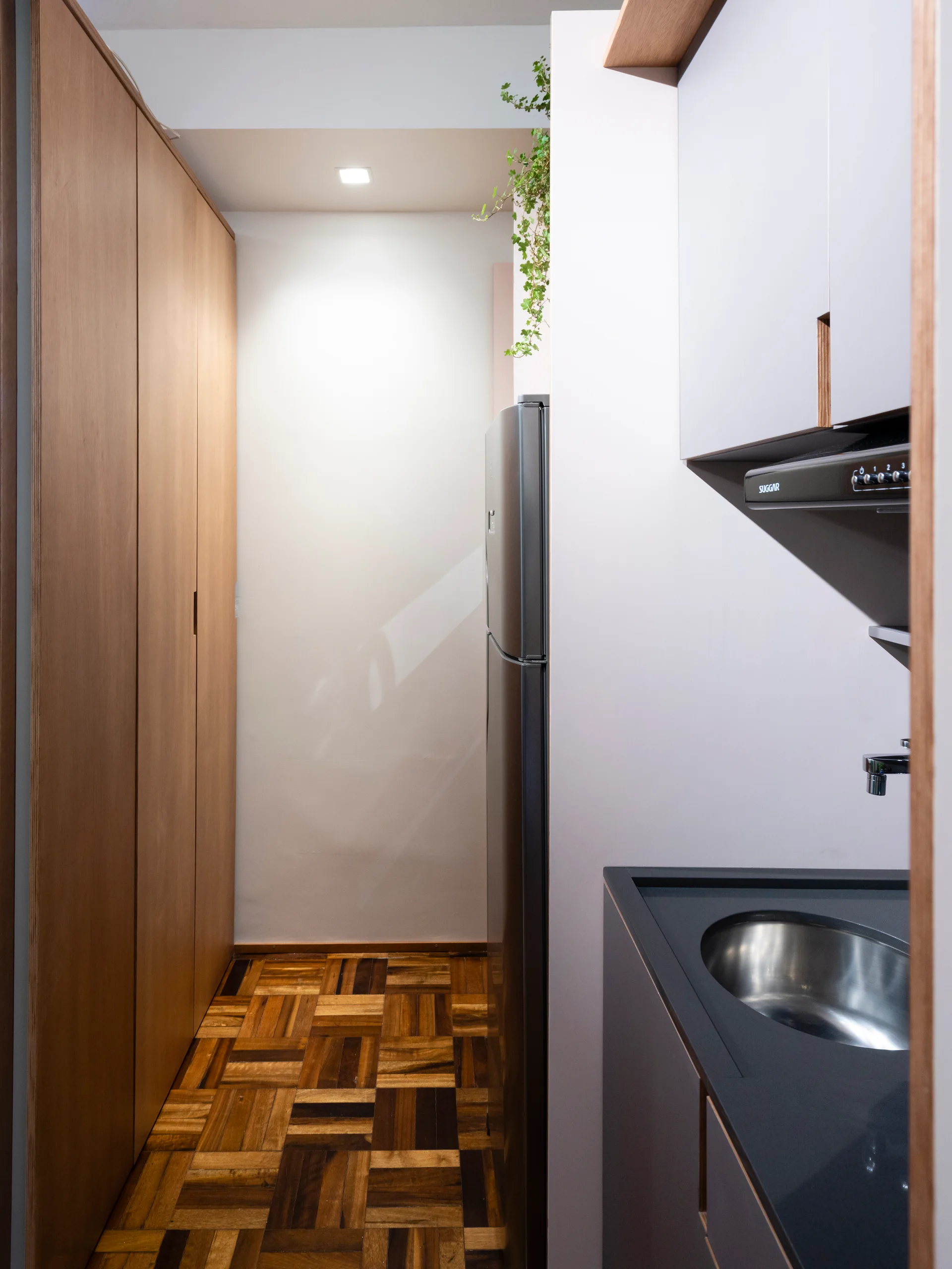 A narrow galley kitchen featuring parquet wood flooring, a tall wooden storage cabinet, and a dark countertop with a recessed stainless steel sink.