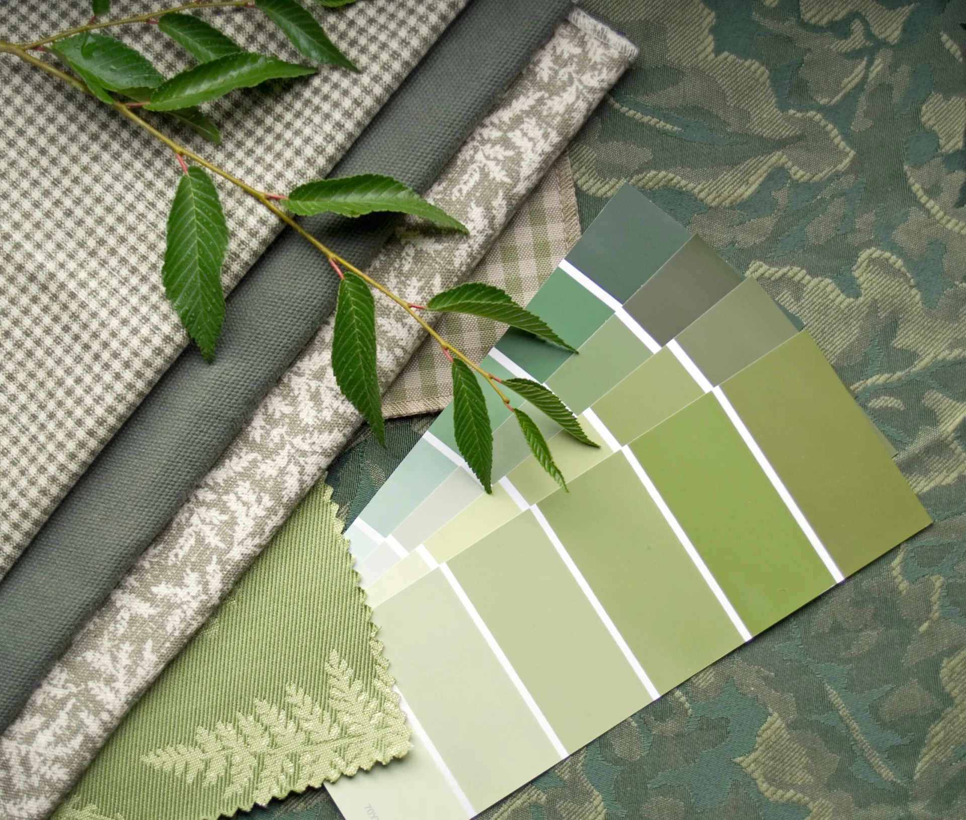 Fabric swatches in various green shades including houndstooth and botanical patterns are layered beneath paint color samples and a green leafy stem.