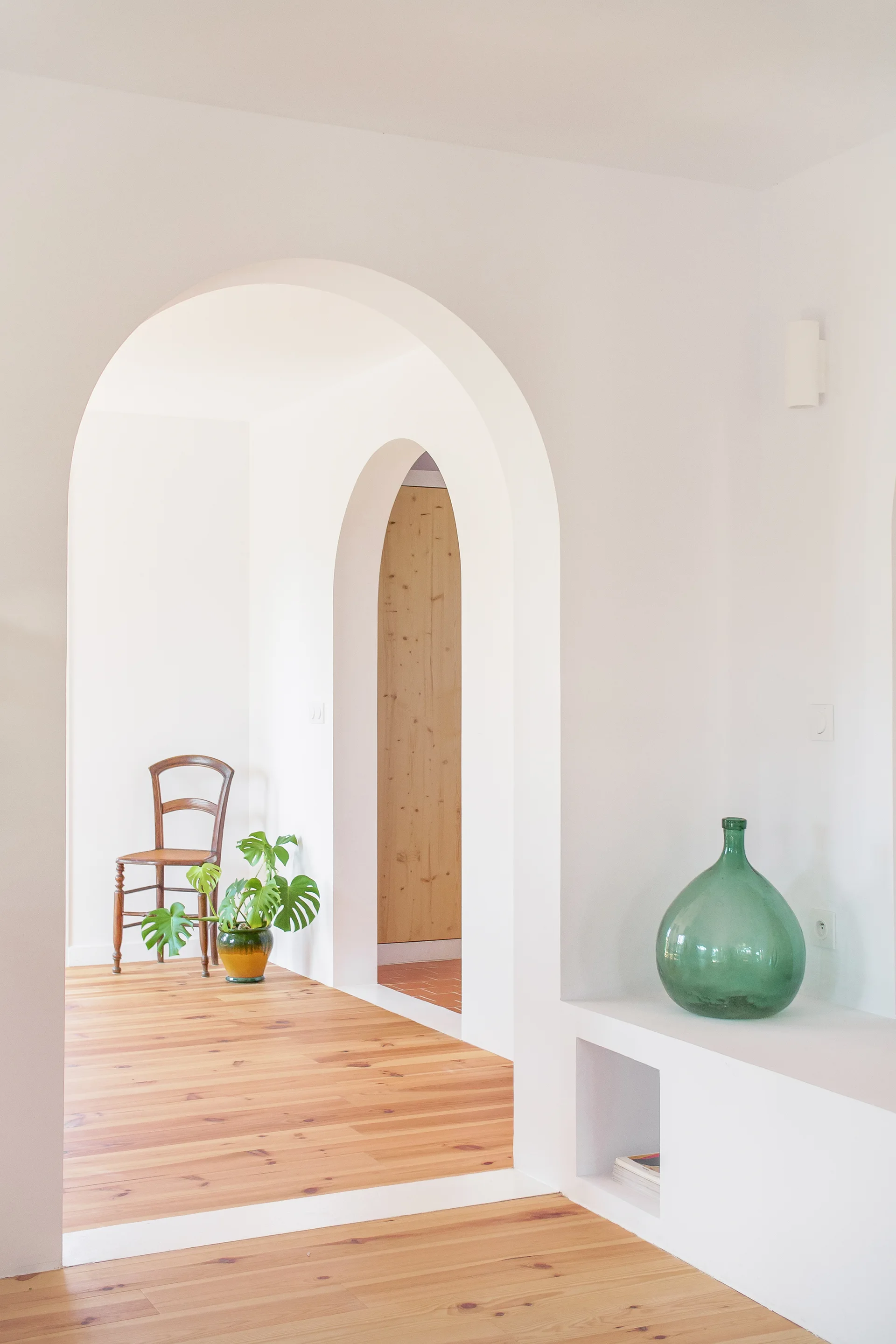A bright white hallway featuring multiple arched doorways, light wood floors, a large green glass vase on a low shelf, and a wooden chair in the background.