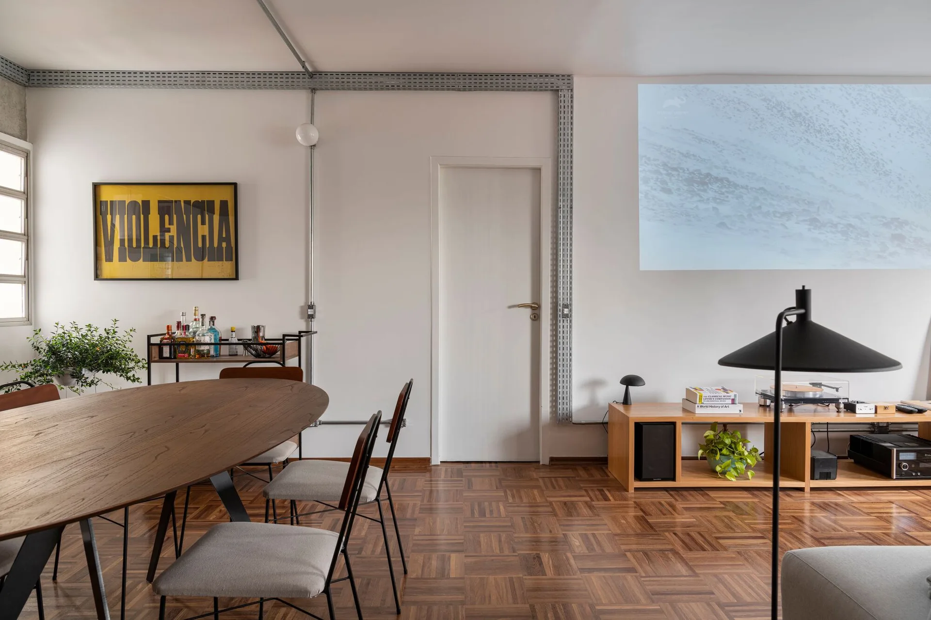 A modern dining room features an oval wood table with grey chairs, exposed metal wall conduits, and a low wooden media console.