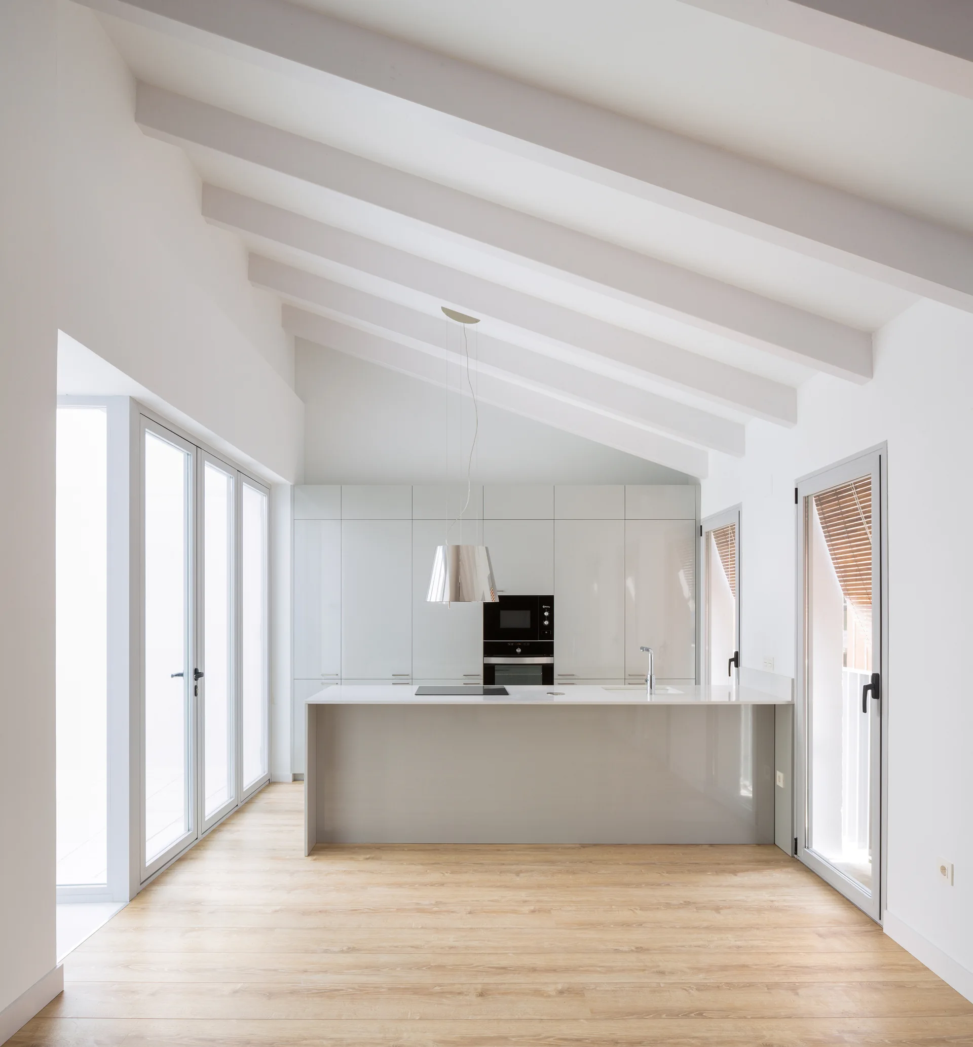 A modern minimalist kitchen with a grey central island, seamless light grey cabinets, and a high vaulted ceiling with white beams.