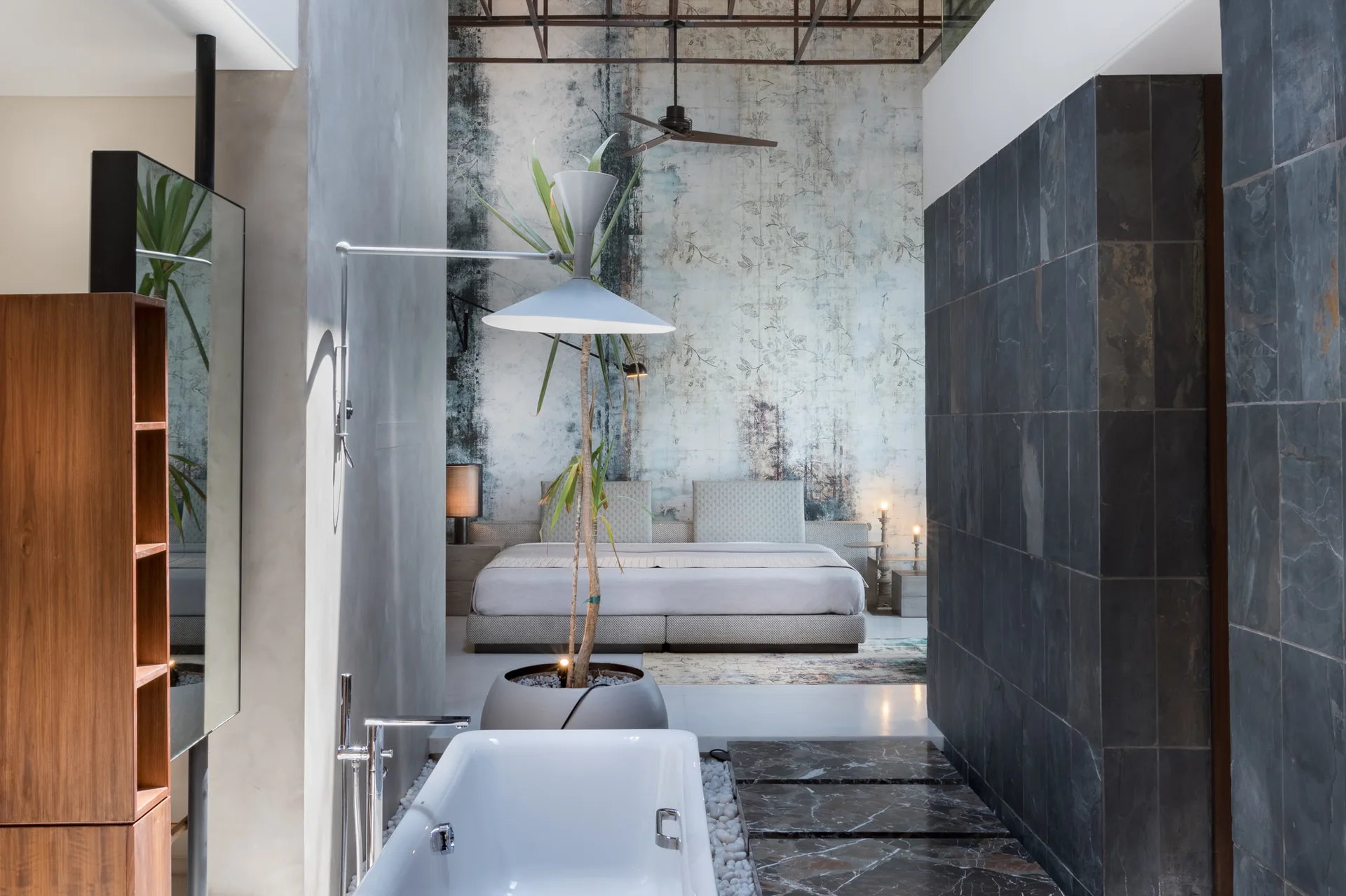 An industrial open-concept bedroom suite features a freestanding white bathtub in the foreground and a bed against a distressed floral accent wall.
