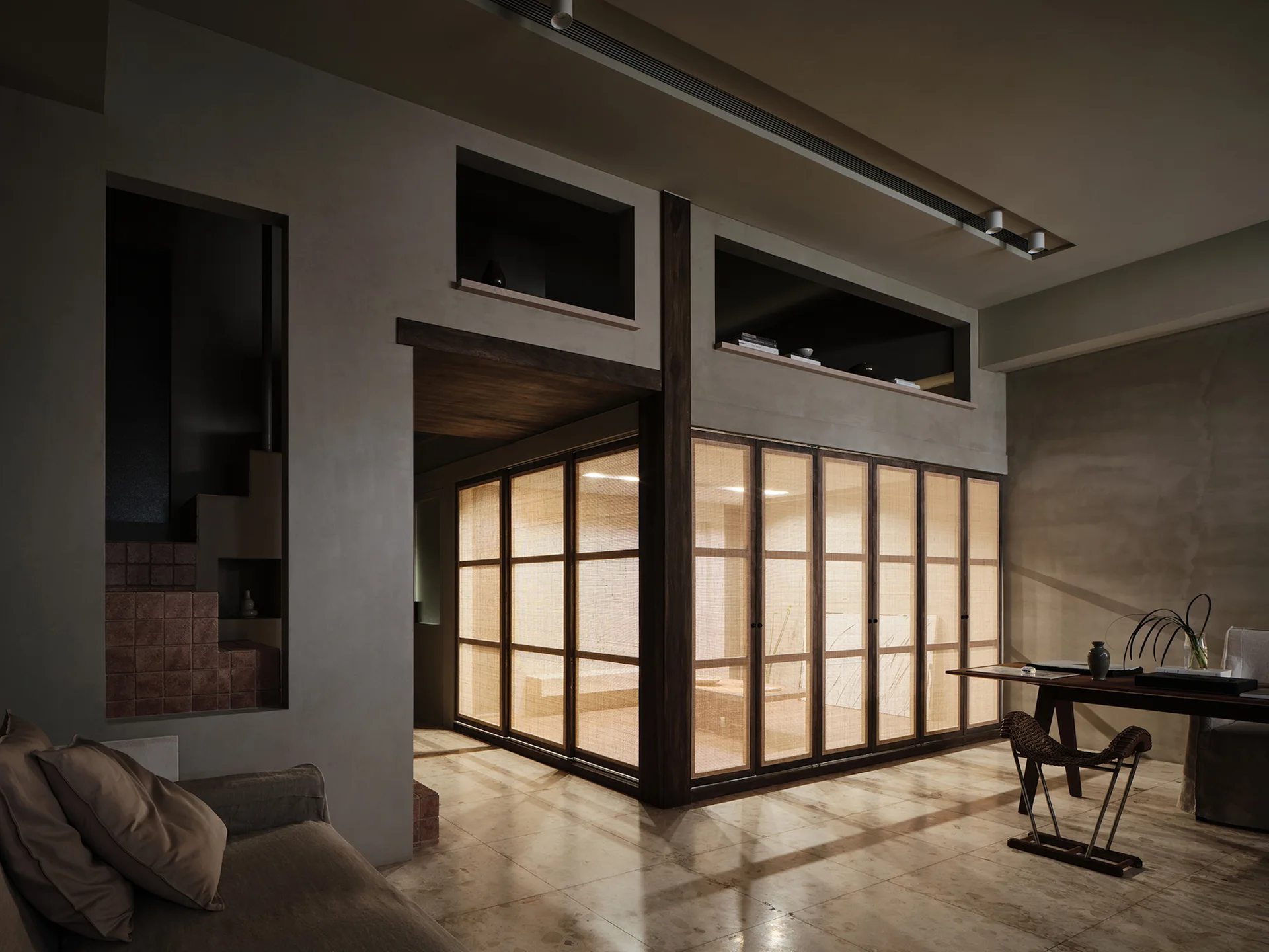 A dimly lit room featuring a large backlit rattan sliding screen partition, concrete-look plaster walls, and beige stone floor tiles.