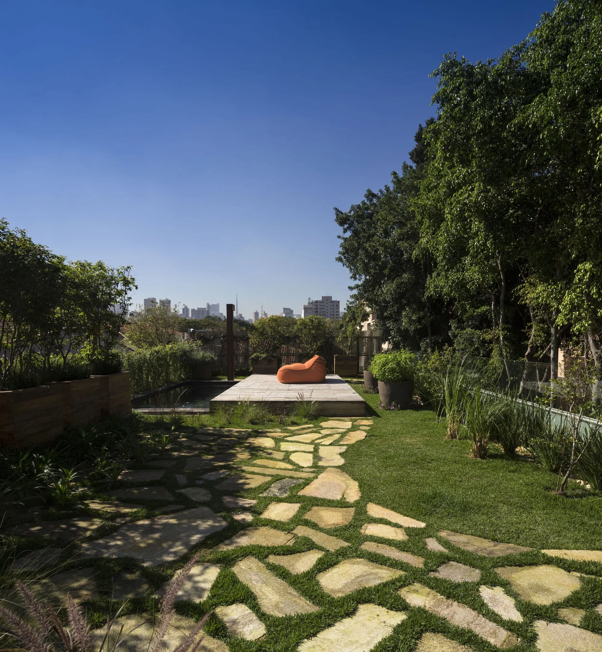 A rooftop garden features a winding flagstone pathway in the grass leading to a wooden deck with an orange lounge chair next to a pool.