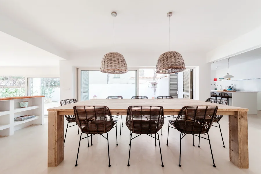 A bright dining room features a large light-wood table surrounded by dark woven rattan chairs beneath two matching woven pendant lamps.