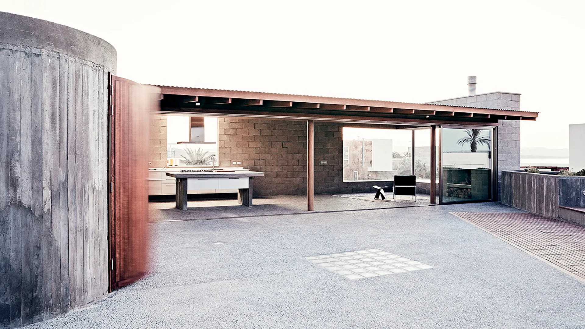A minimalist outdoor patio features concrete block walls, exposed wood ceiling beams, a large sliding glass door, and an open-air kitchen island.
