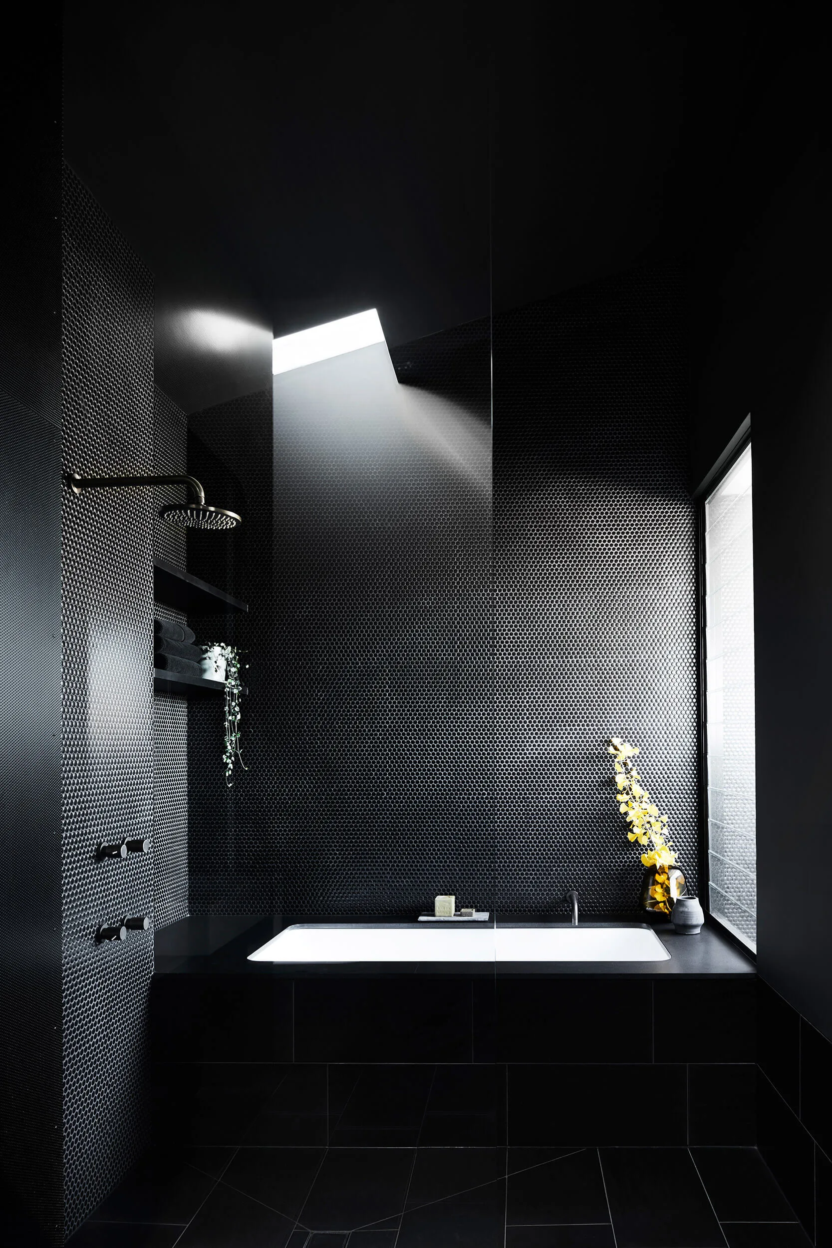 A modern bathroom features black penny round tiles, a white built-in bathtub, and a stream of natural light from an overhead skylight.