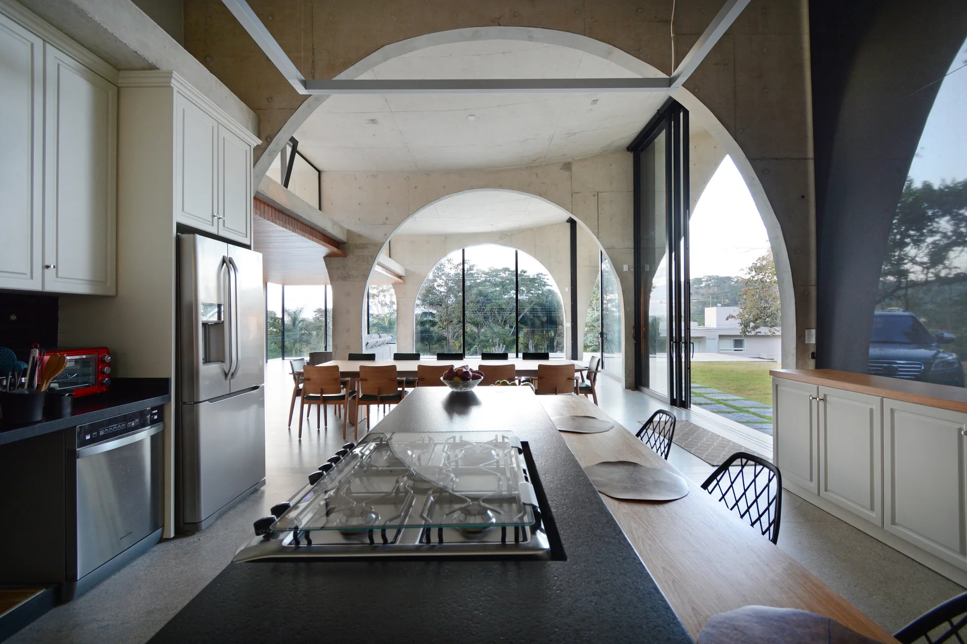 A modern kitchen with a grey countertop island sits under large raw concrete arches framing a dining table and garden views.