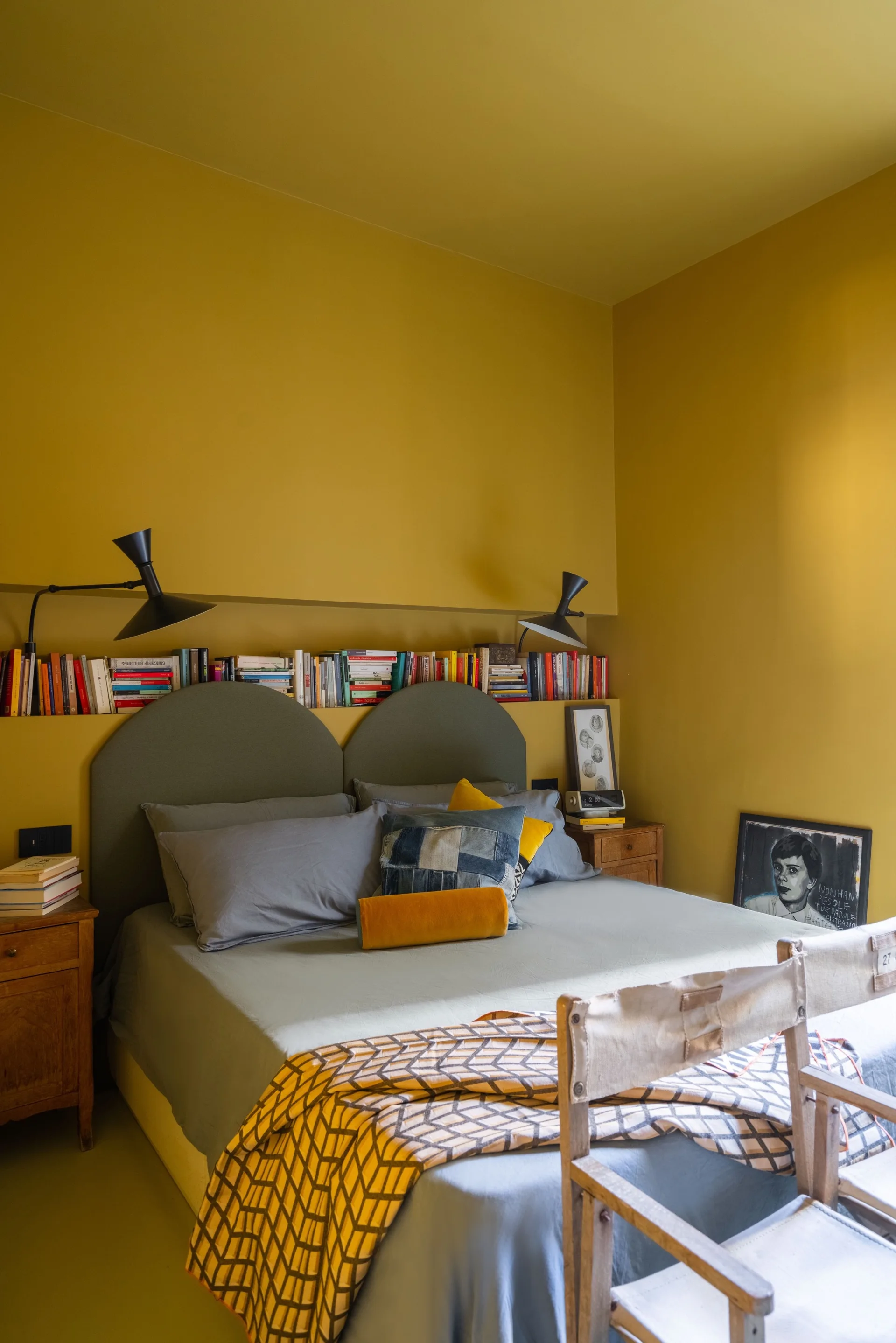 A mustard yellow bedroom features a bed with a green arched headboard and a recessed wall shelf filled with books under two black sconces.