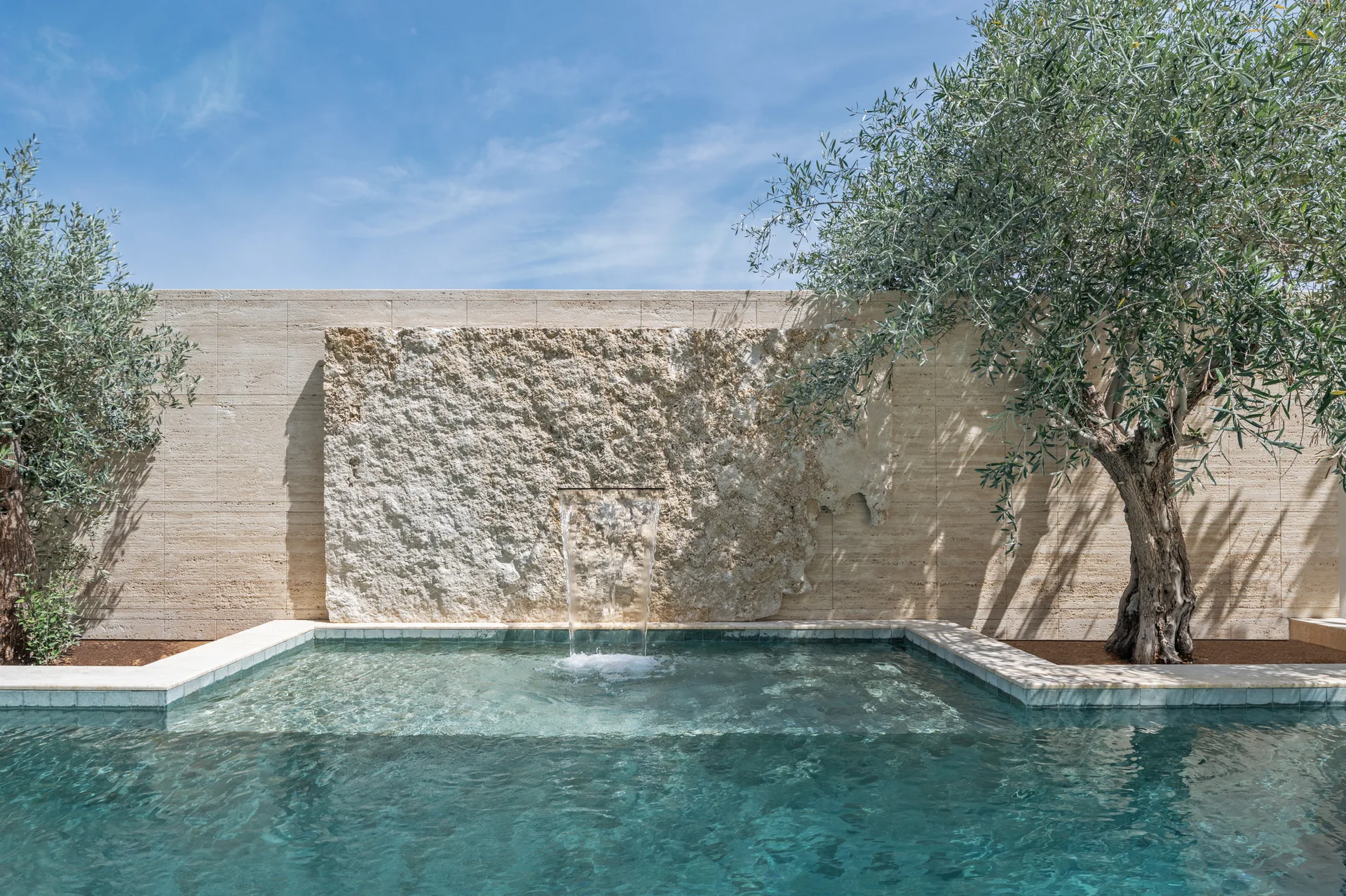 A turquoise swimming pool features a smooth travertine wall with a rough stone water feature and cascading waterfall next to an olive tree.