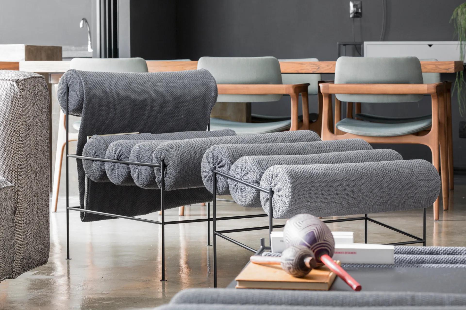 A contemporary gray lounge chair with cylindrical bolster cushions sits on a polished floor in front of a wooden dining table set.
