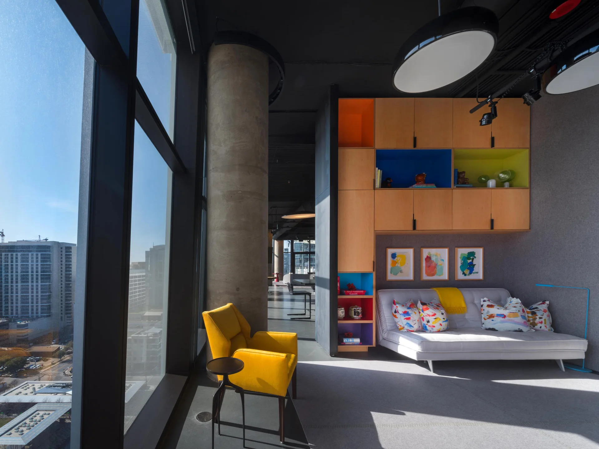 A modern living room features a grey sofa beneath wood cabinetry with colorful niches, an exposed concrete column, and a yellow armchair by a window.