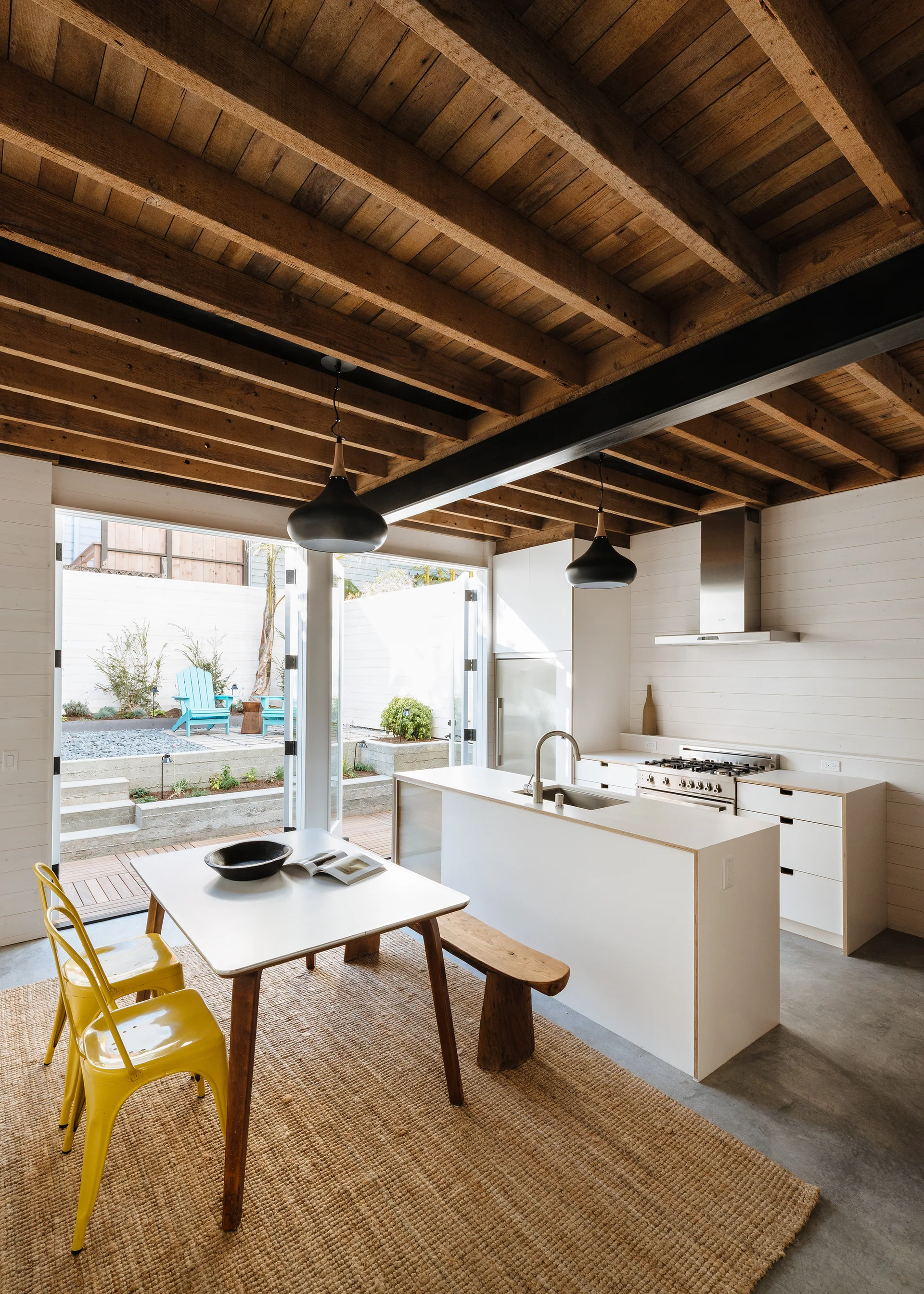 An open-concept kitchen and dining area features an exposed wood ceiling, a white island, a dining table with yellow chairs, and concrete flooring.