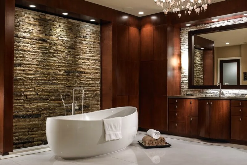 A modern bathroom features a white oval freestanding bathtub set against a textured stone wall and dark wood cabinetry with a large mirror.