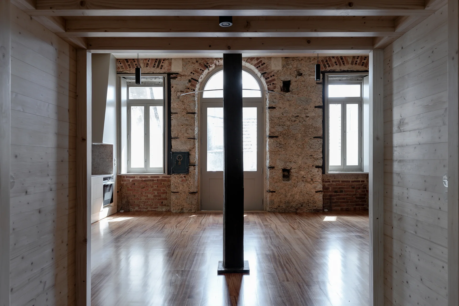 An empty renovated room features a glossy wood floor, a central dark column, and an exposed stone wall with tall windows.