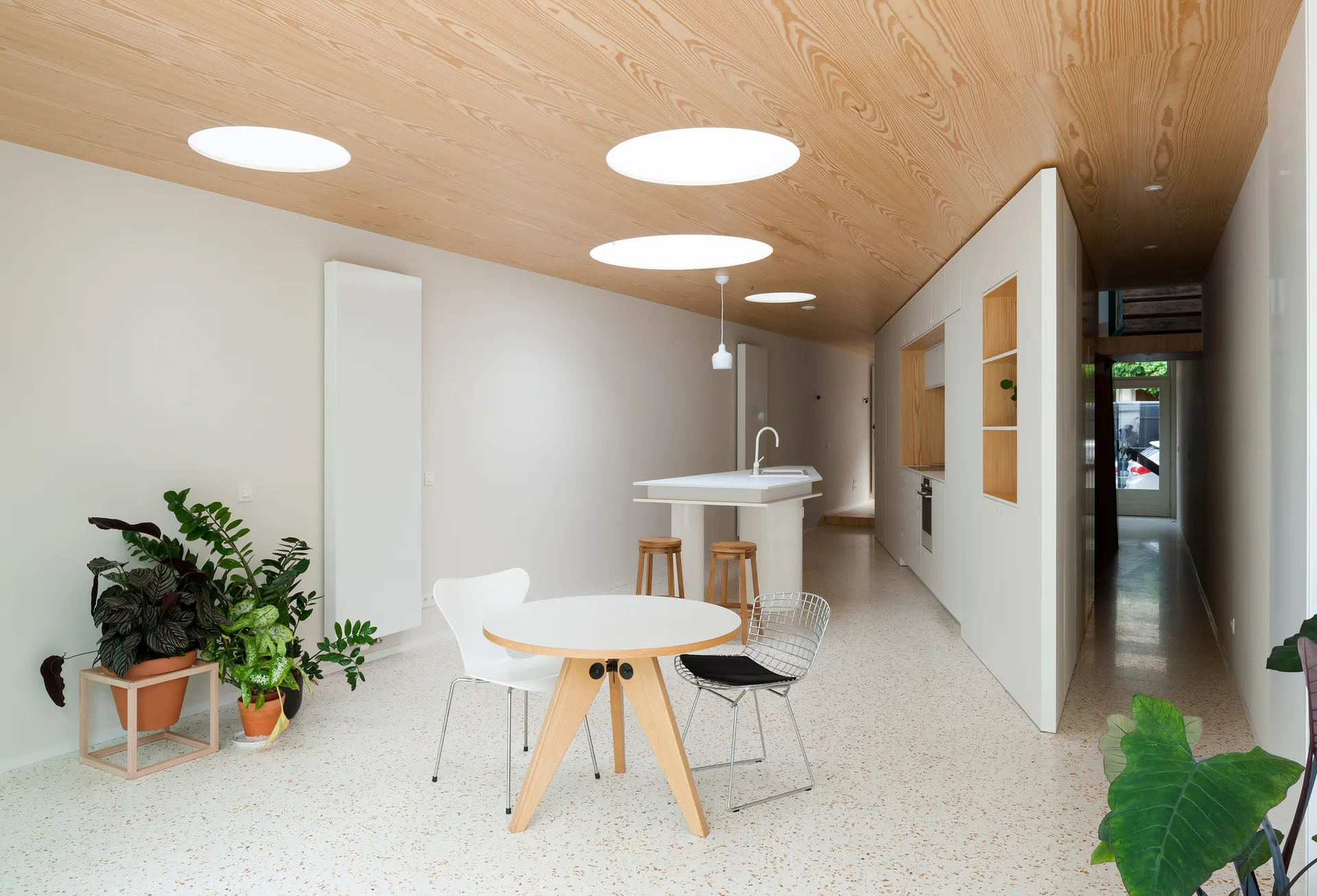 A minimalist kitchen and dining area with a wood-veneered ceiling, circular skylights, terrazzo-style floors, and a round wooden dining table.