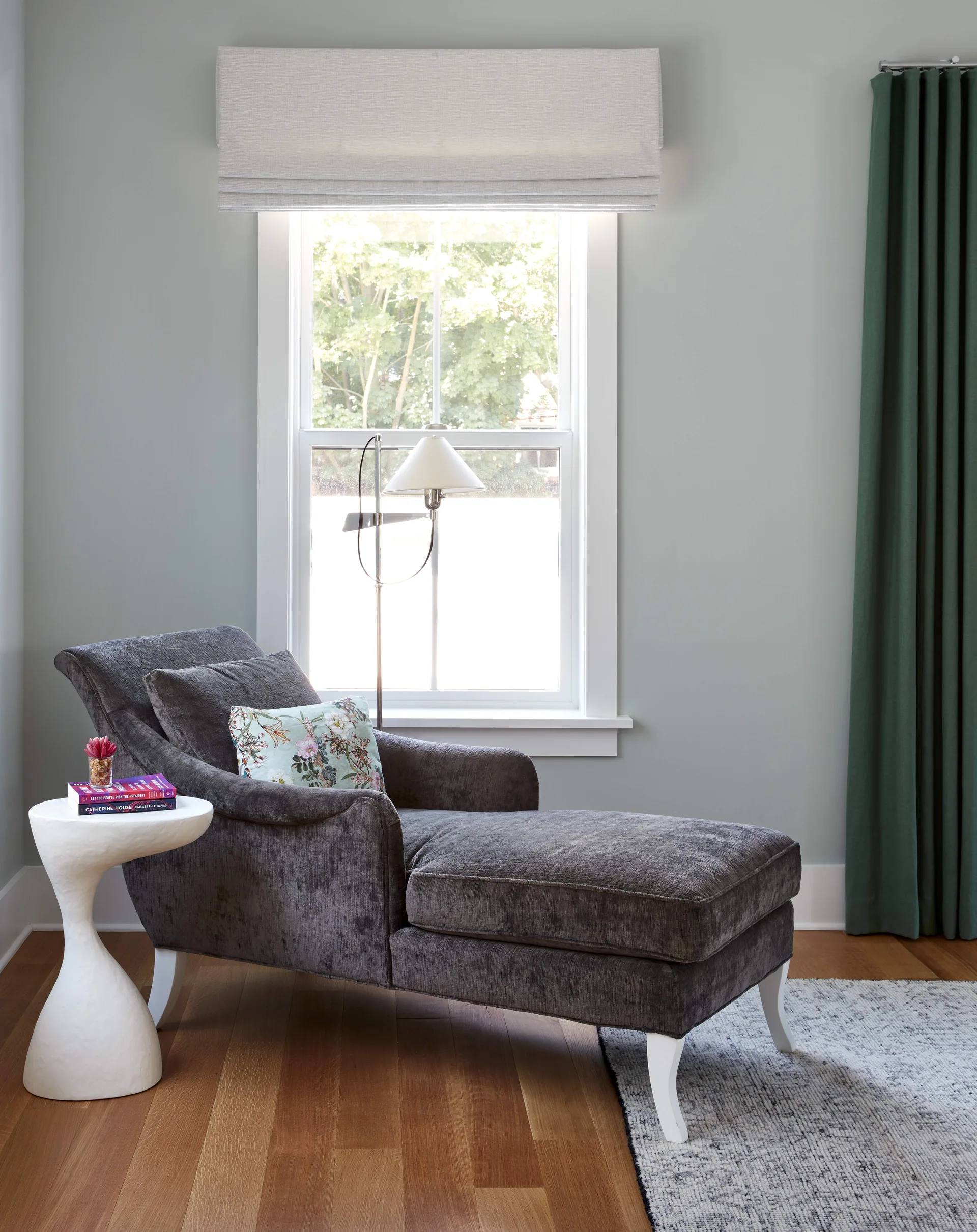 A charcoal chaise lounge with white legs sits in a gray reading nook next to a white hourglass side table and a window with green drapery.