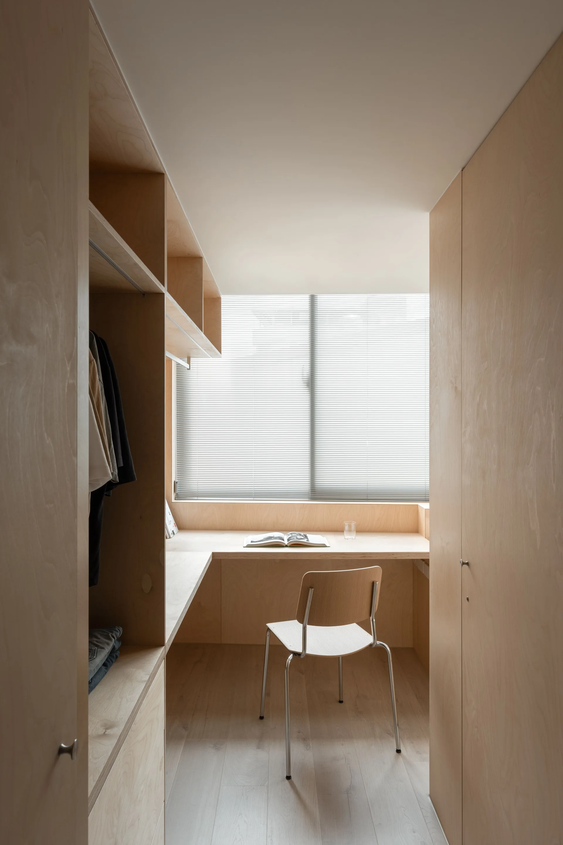 A minimalist home office workspace features a built-in plywood desk beneath a window with blinds, flanked by custom wood closets.