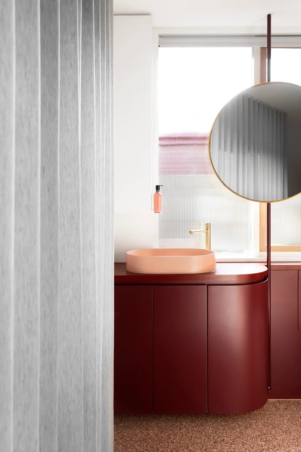 A modern bathroom with a curved deep red vanity cabinet, a peach vessel sink, a gold faucet, and a circular mirror on a vertical pole.