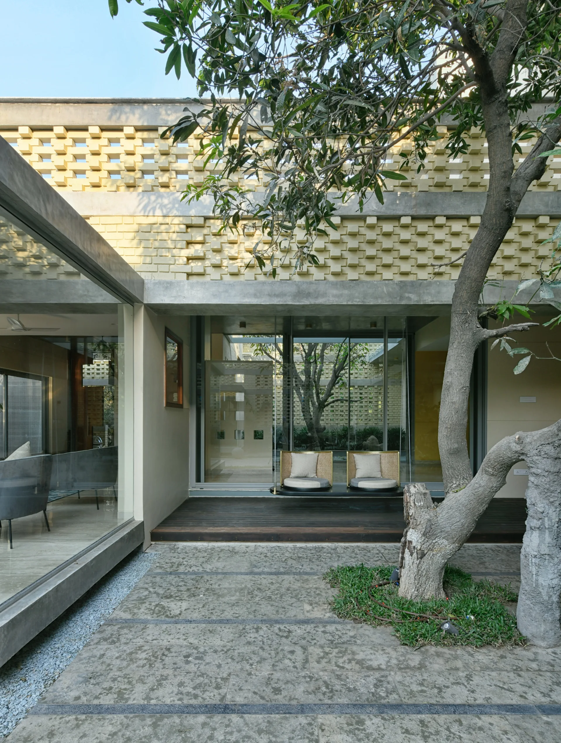 A modern concrete patio features a dark wood deck with two hanging chairs in front of a glass window and a textured yellow brick screen wall.