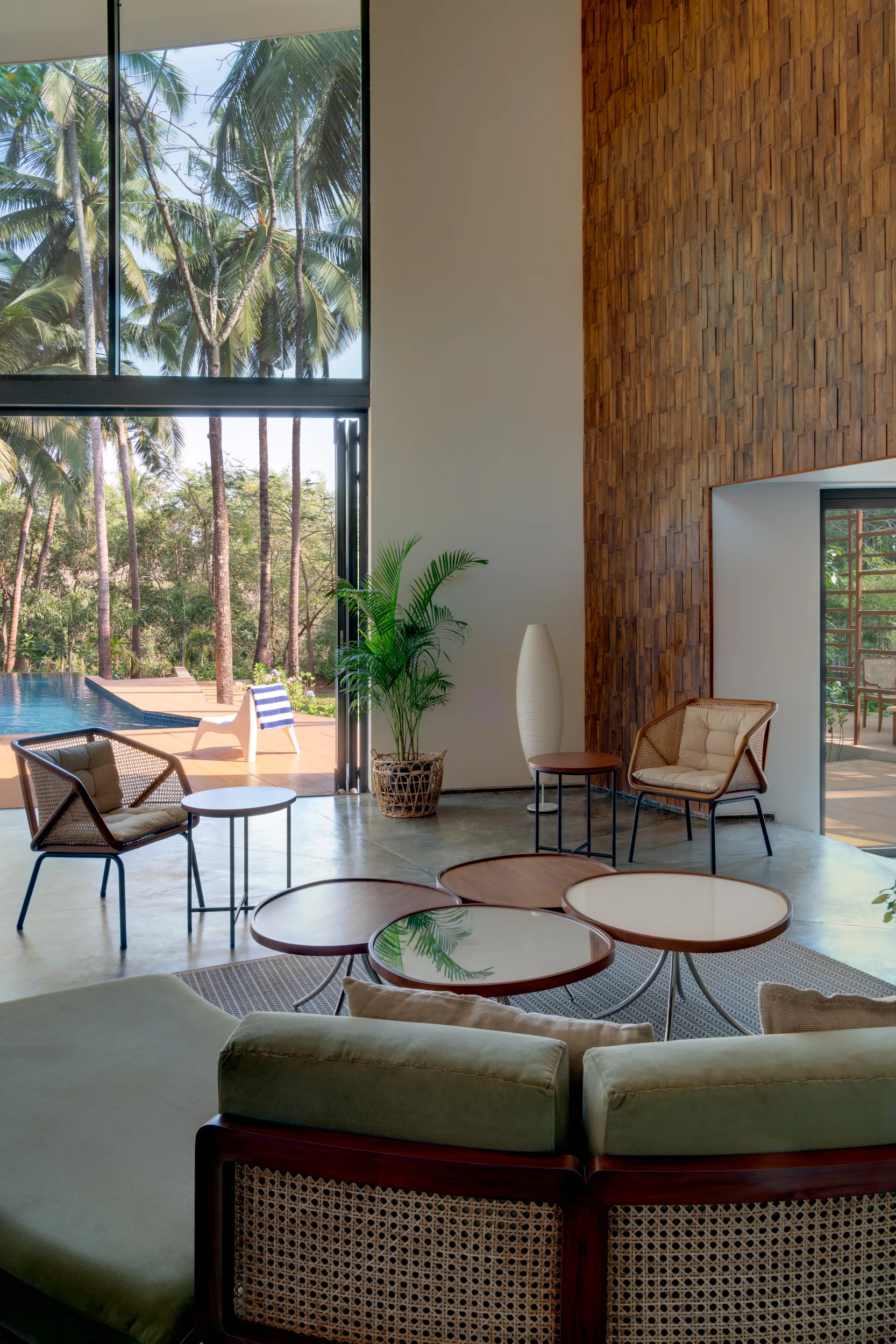 A modern tropical living room with rattan armchairs, circular coffee tables, a tall textured wood wall, and glass doors opening to a pool.